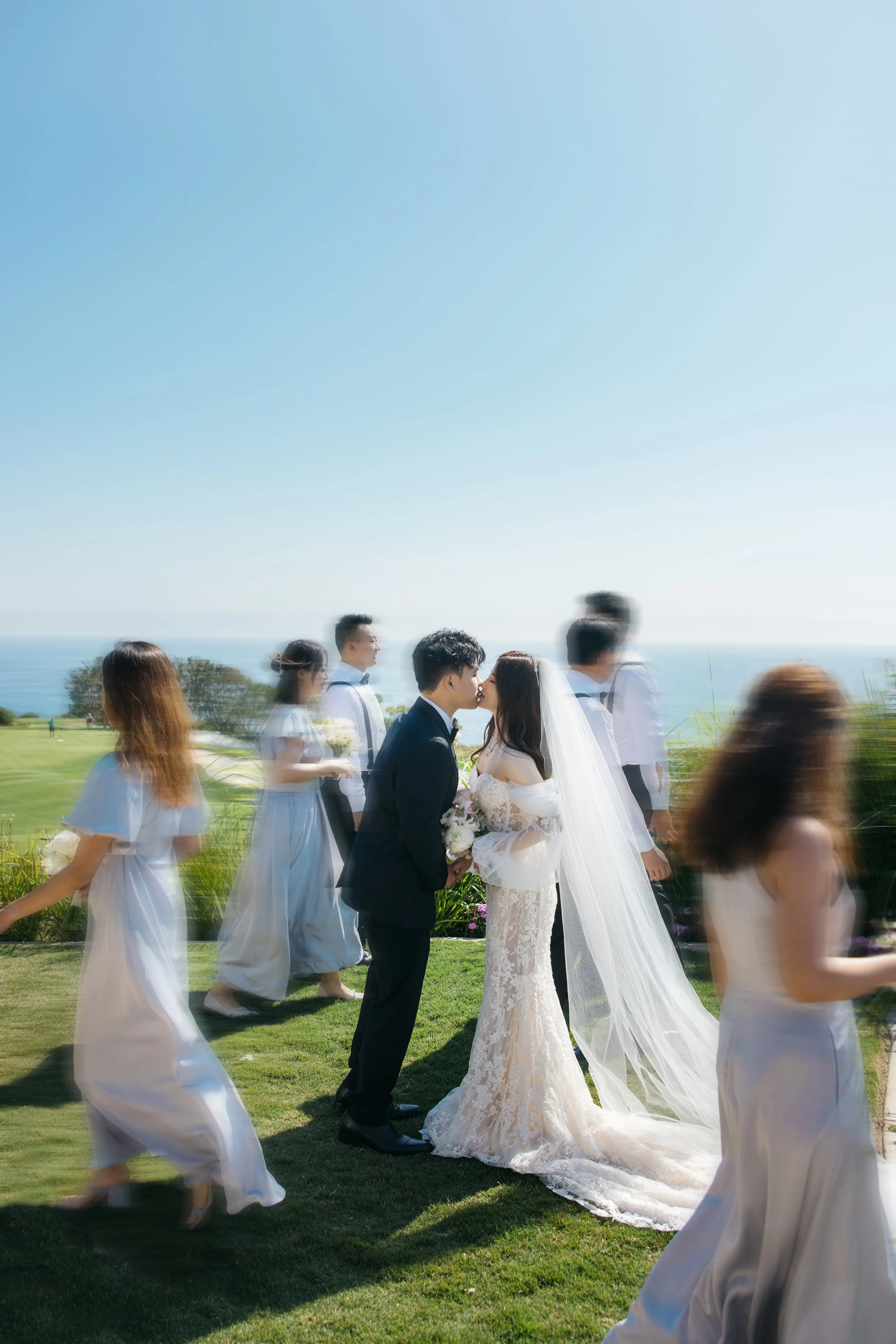 A bride and groom sharing a kiss during their outdoor wedding ceremony with friends and family in the background, overlooking the ocean on a bright, clear day.