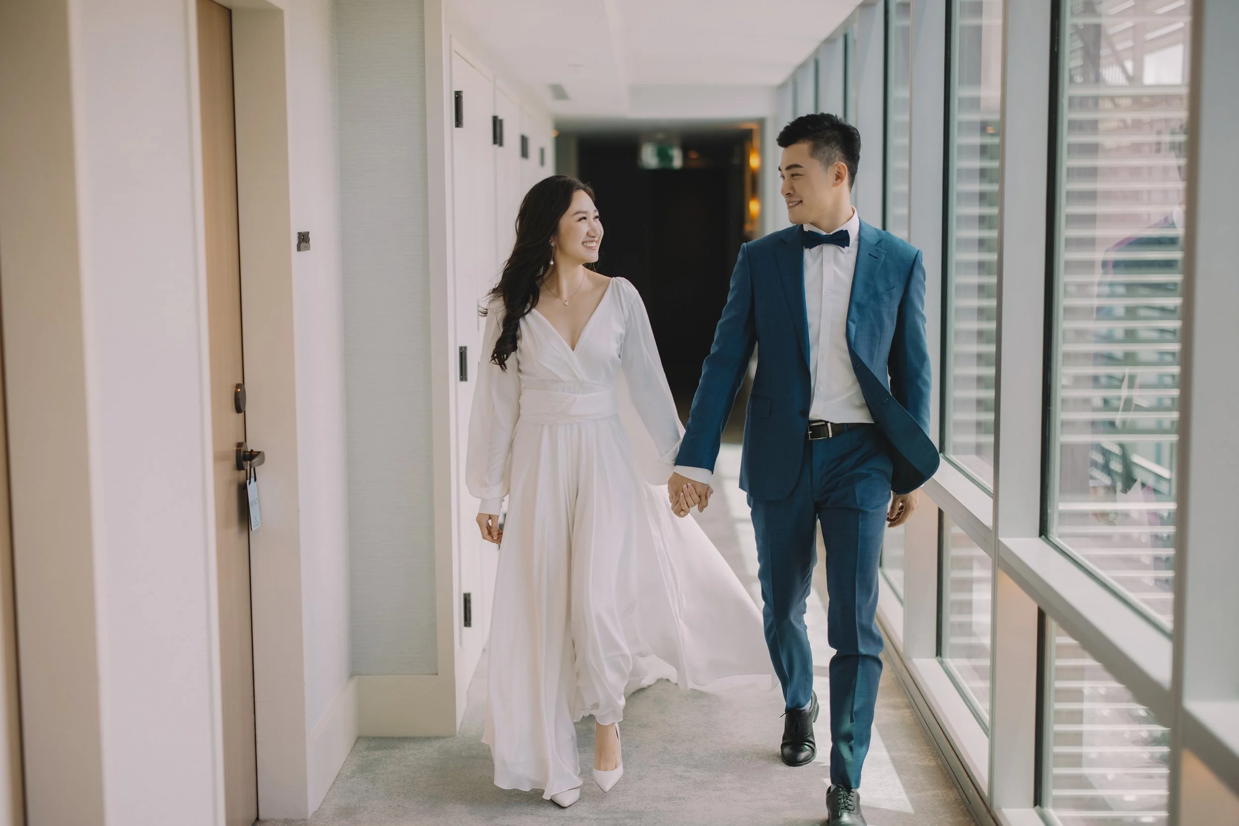 A happy couple walking hand in hand down a hallway with large windows, the woman in a white dress and the man in a blue suit and bow tie, smiling at each other.