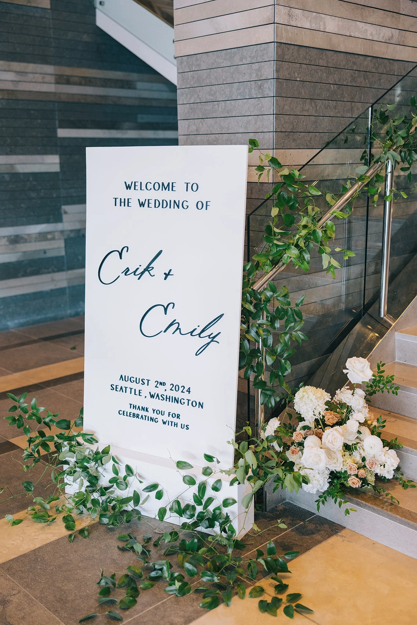 Wedding welcome sign with greenery and flowers at the base, placed next to a staircase in an indoor venue. The sign has the names Crik and Emily, the date August 2nd, 2024, and the location Seattle, Washington, along with a thank you message.