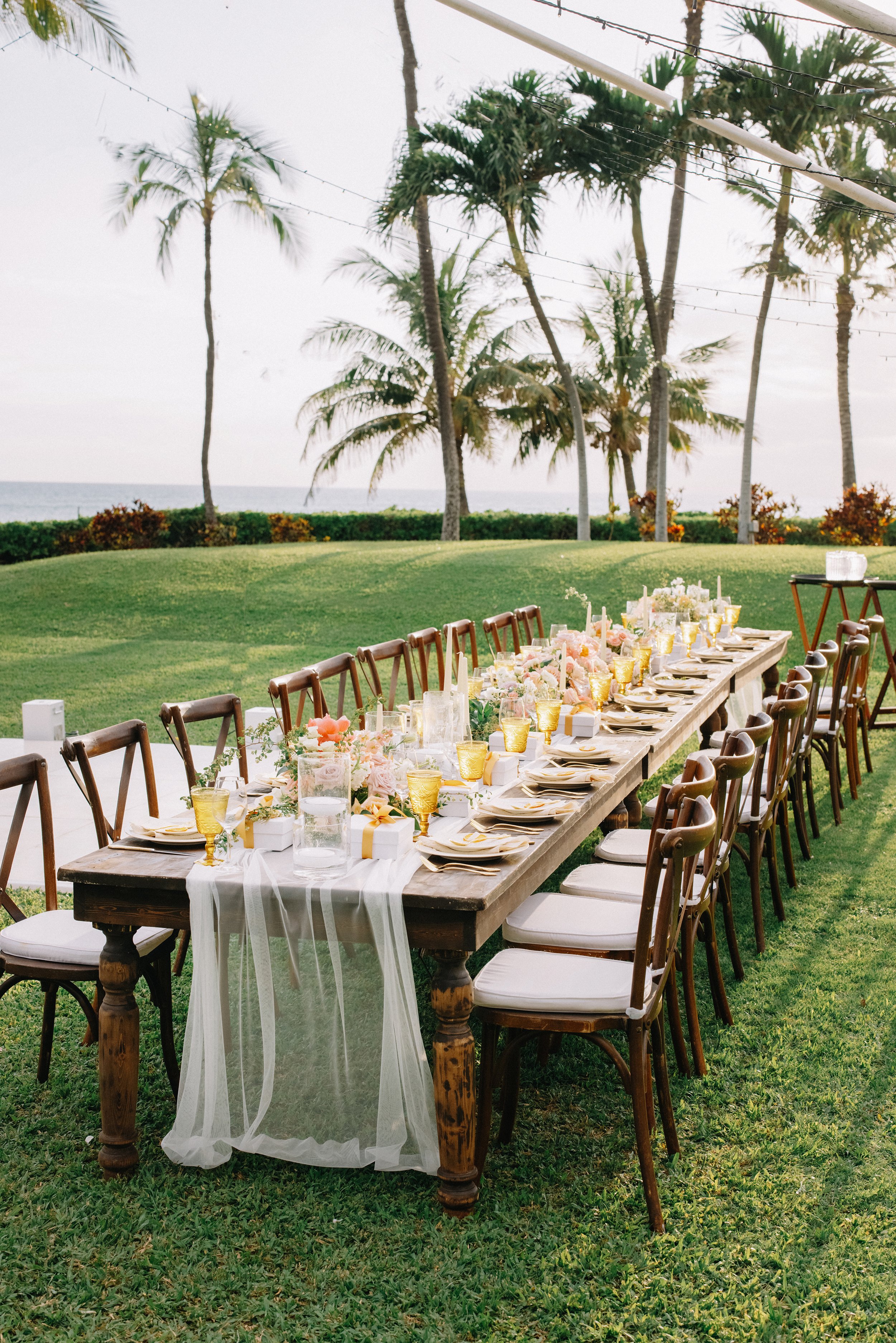 Outdoor wedding reception table setup with floral arrangements, candles, and gift boxes, set on a grassy lawn with palm trees and ocean in the background.