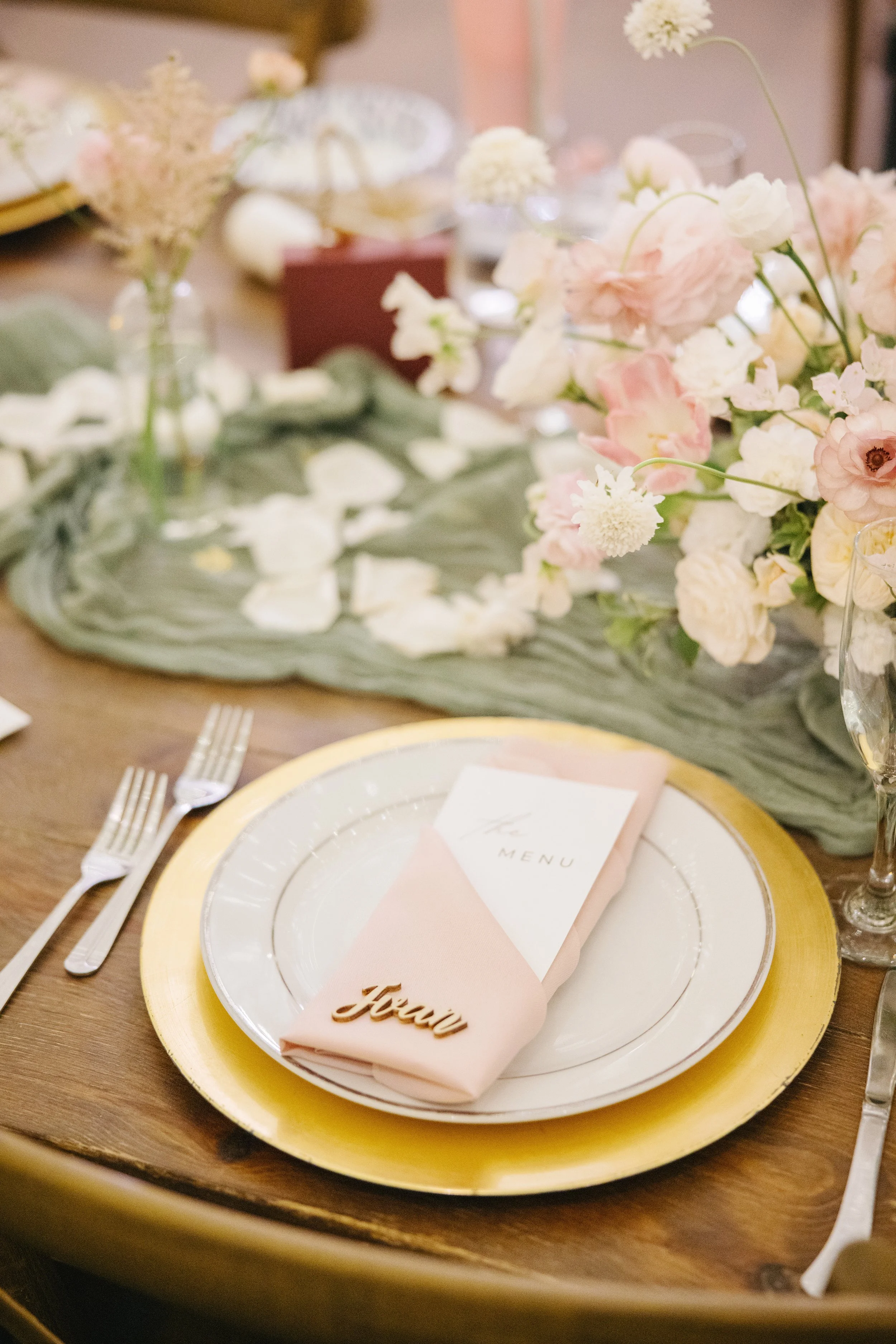 Table setting with a white plate, gold charger, pink napkin with a wooden 'fleur' label, and a menu, surrounded by floral decorations and glassware.