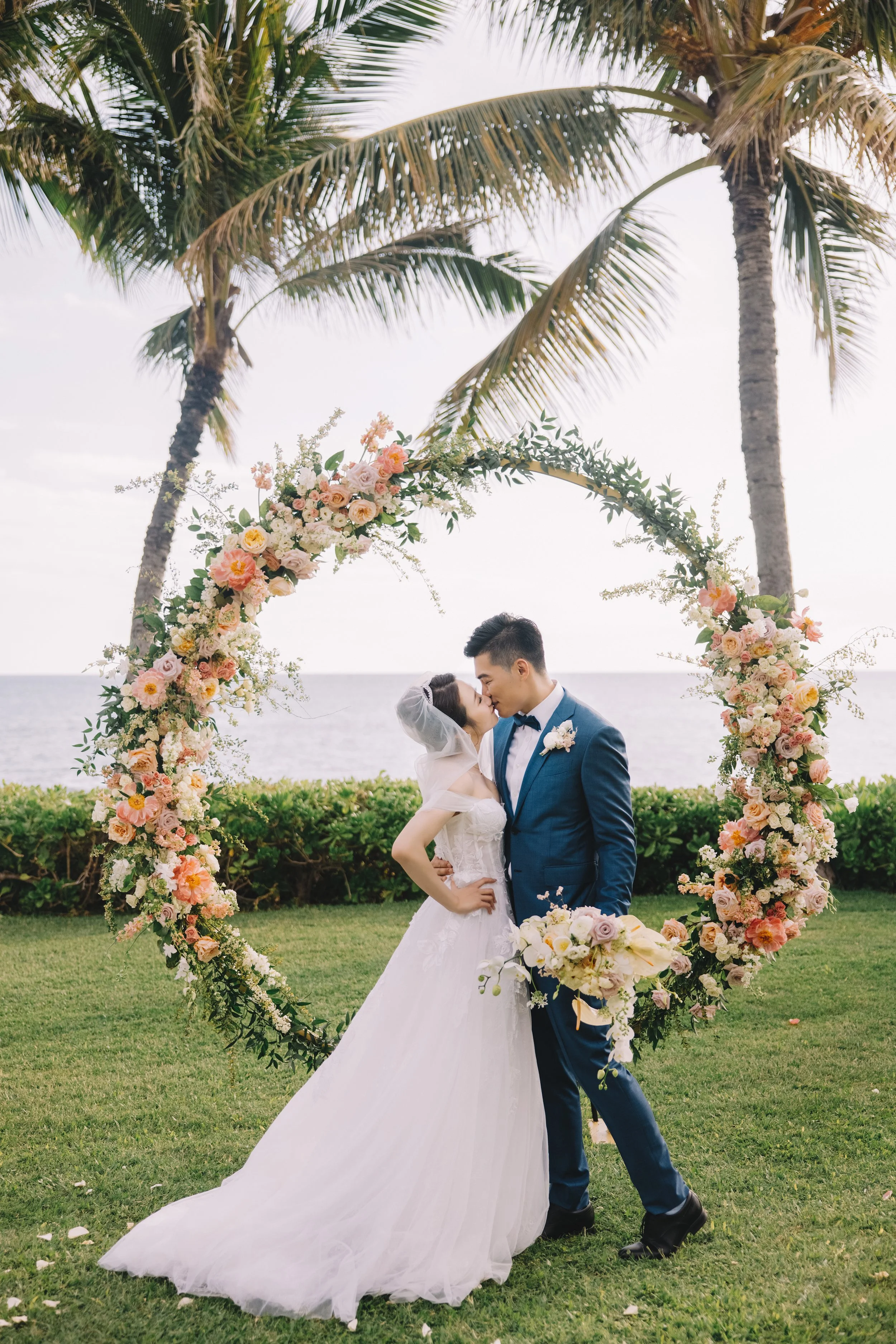 A bride and groom kiss under a heart-shaped floral arch on a grassy area near the ocean, with palm trees overhead.