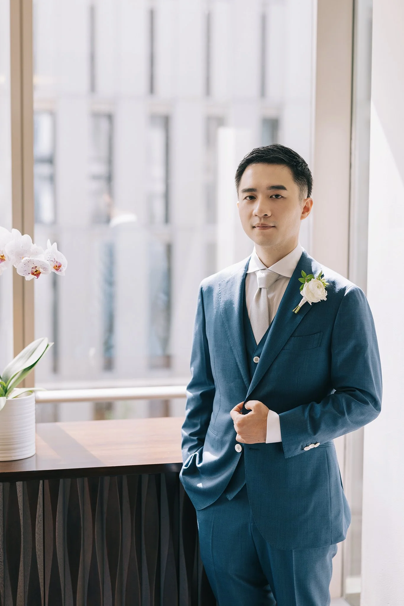 A young man in a blue suit with a white shirt and a matching tie standing indoors near a window, with a white flower corsage on his lapel, next to a wooden table with a white potted orchid.