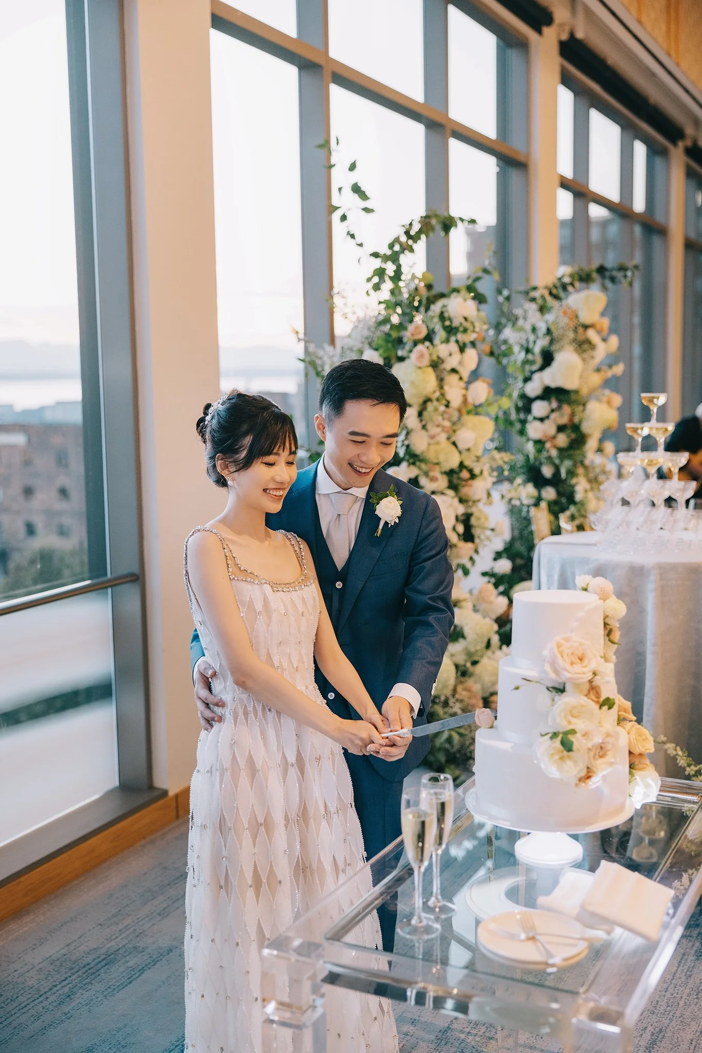 A newlywed couple is cutting their wedding cake together at their reception. They are standing in front of a large flower arrangement with white and light pink flowers, inside a venue with large windows and a city view.