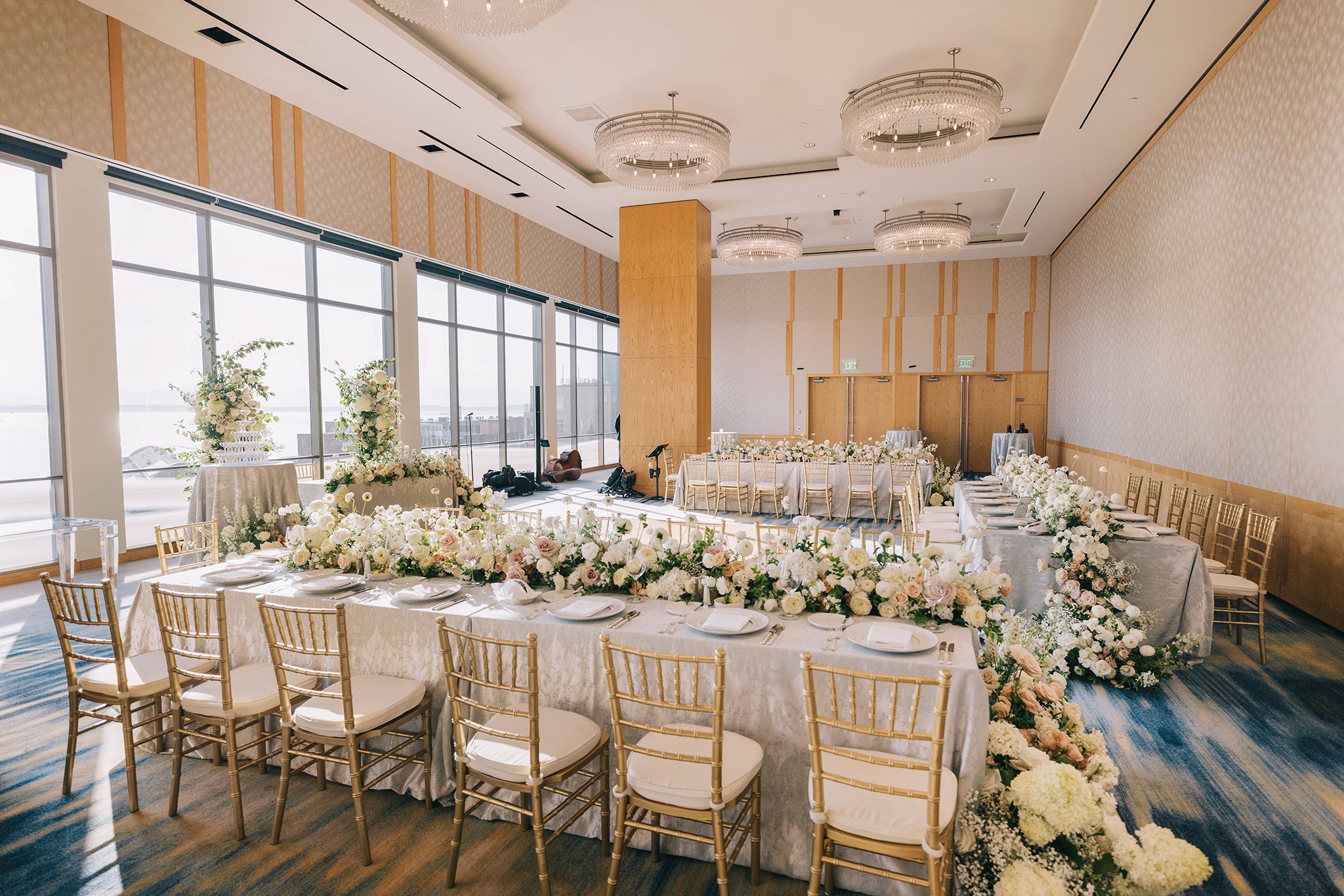 Elegant wedding reception setup with long tables covered in white tablecloths, decorated with white and blush flowers, gold chairs, and large floral arrangements near floor-to-ceiling windows with natural light.