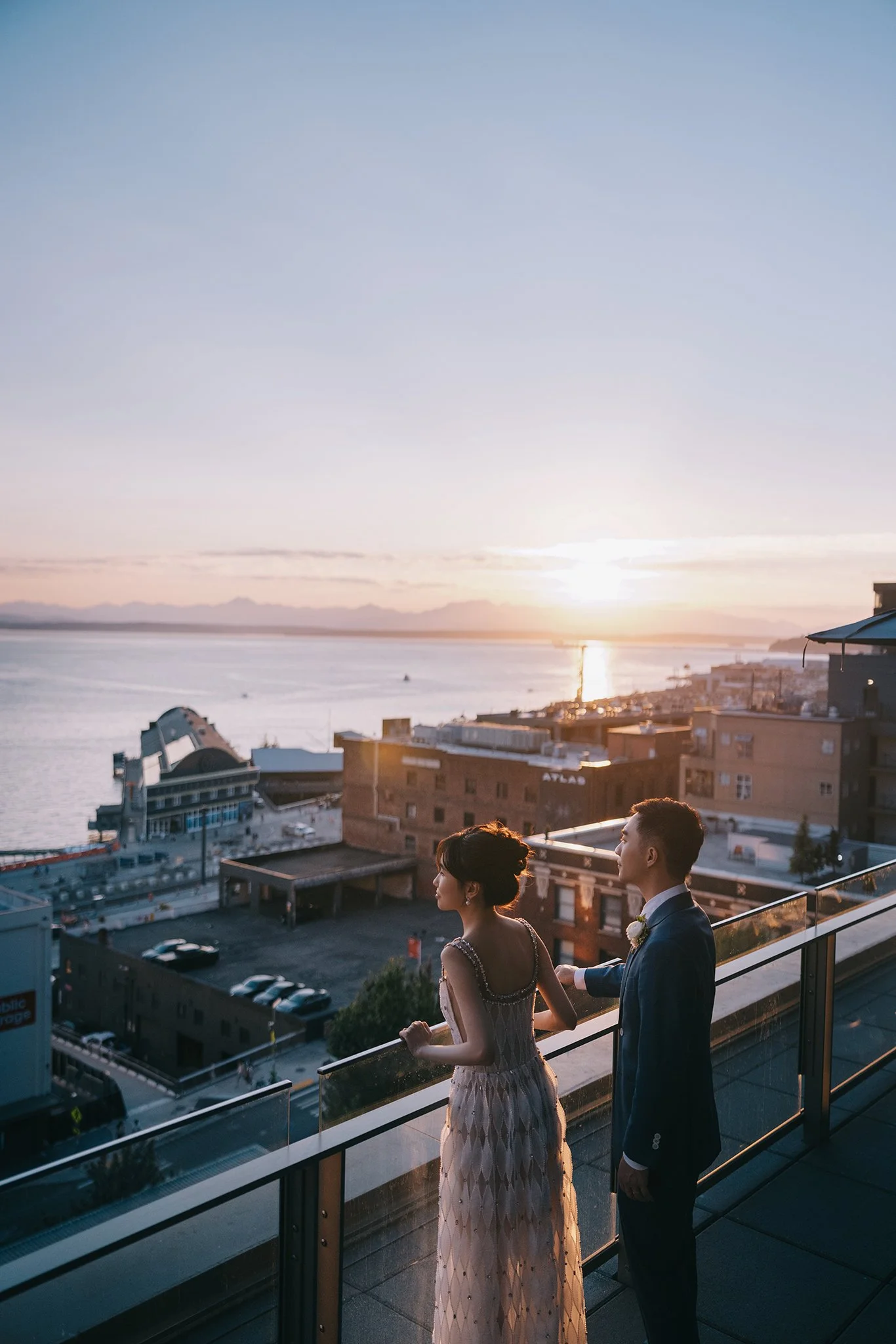 A couple in formal attire standing on a balcony overlooking a city and water during sunset.
