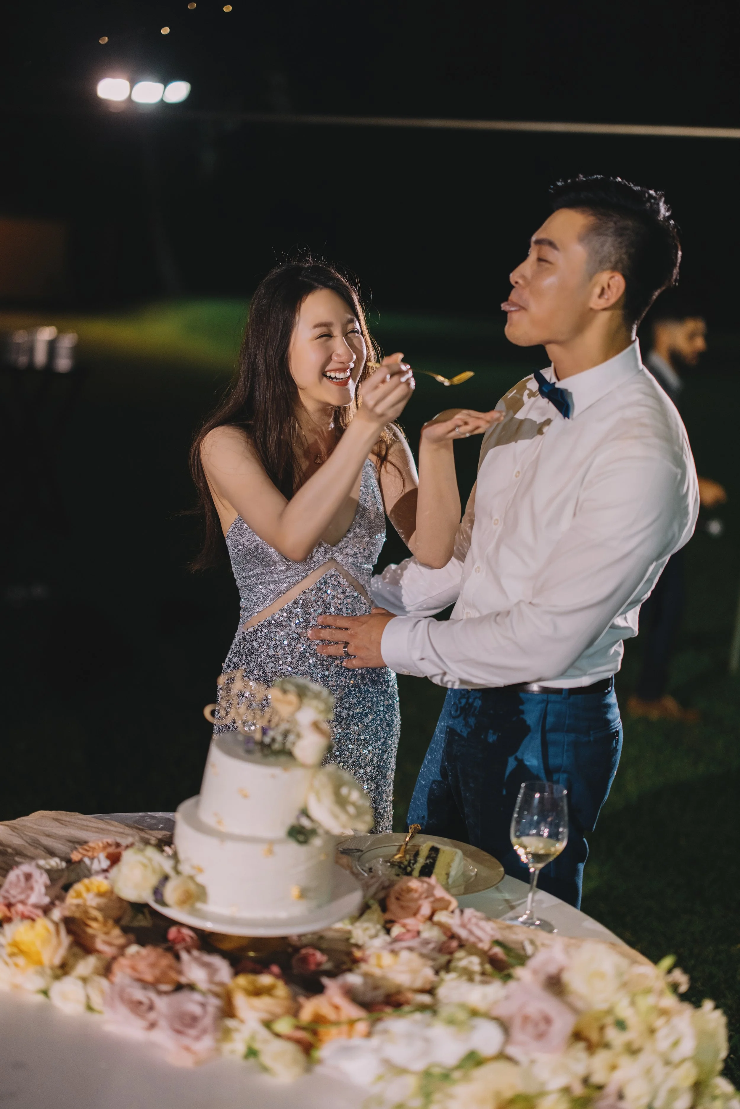 A bride and groom celebrate their wedding reception outdoors at night, with a wedding cake and flowers on the table.