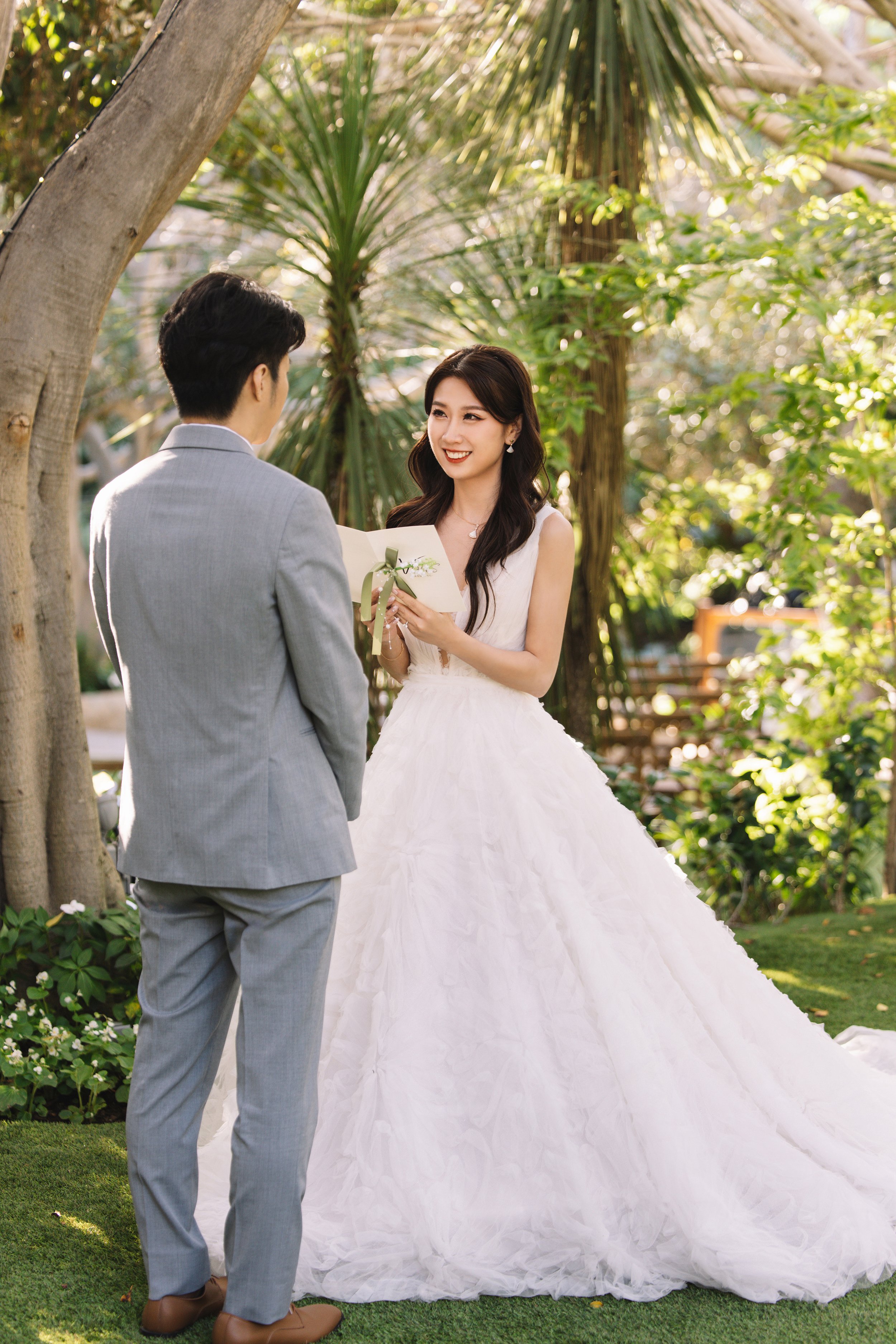 A woman in a white wedding dress is smiling and holding a letter with a ribbon, while a man in a light grey suit is facing her, in a lush garden setting during a wedding ceremony.