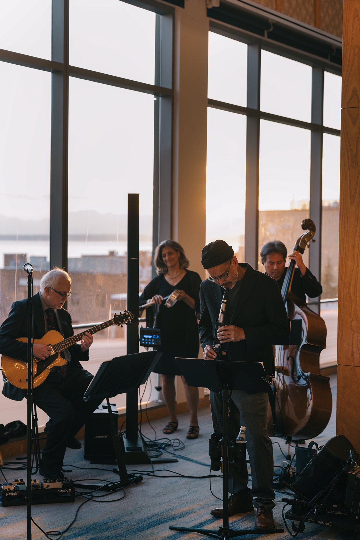 Musicians playing instruments during a live performance in a room with large windows and sunset outside.