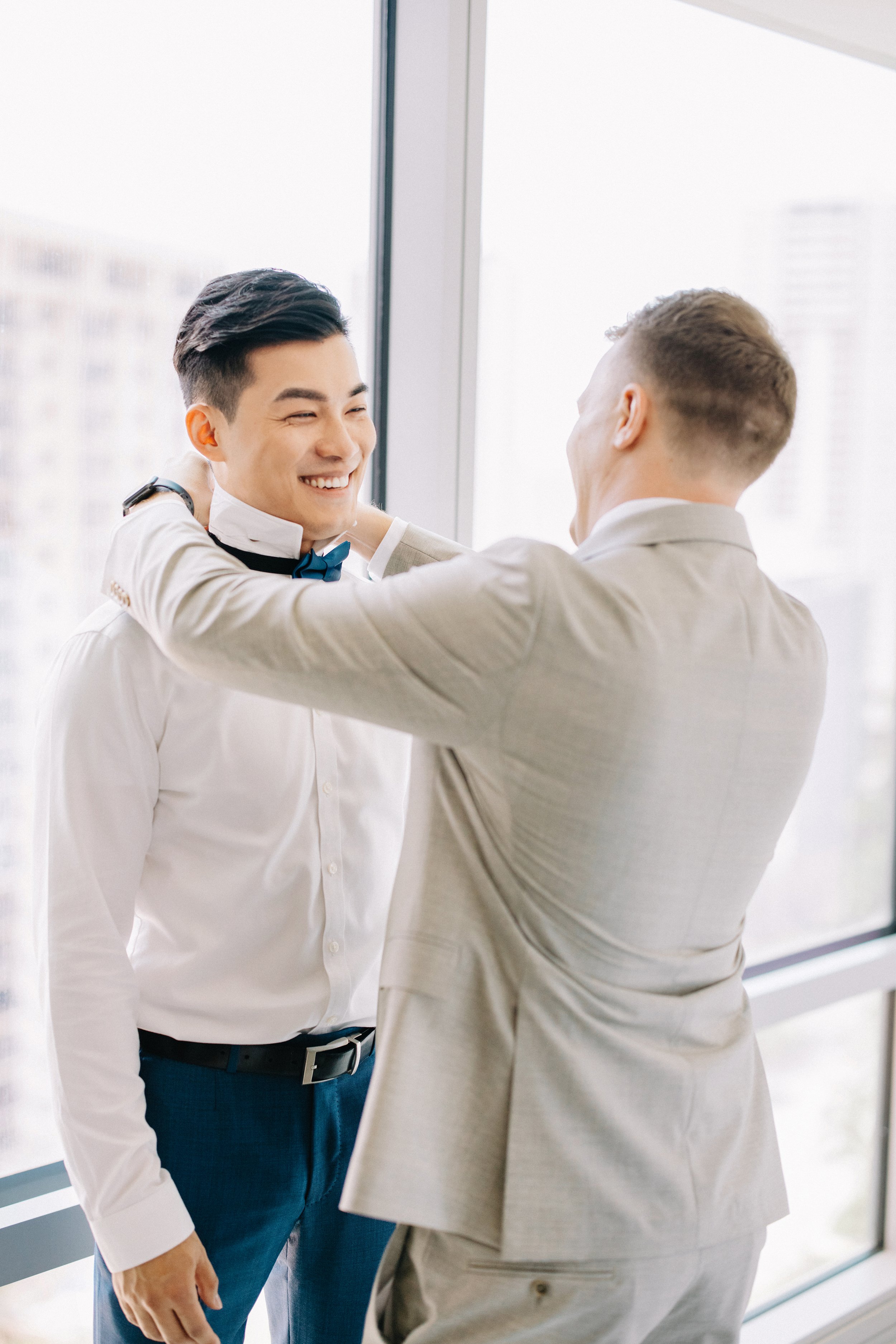 Two men in suits smiling, one helping the other with his bow tie in front of a large window with city buildings outside.