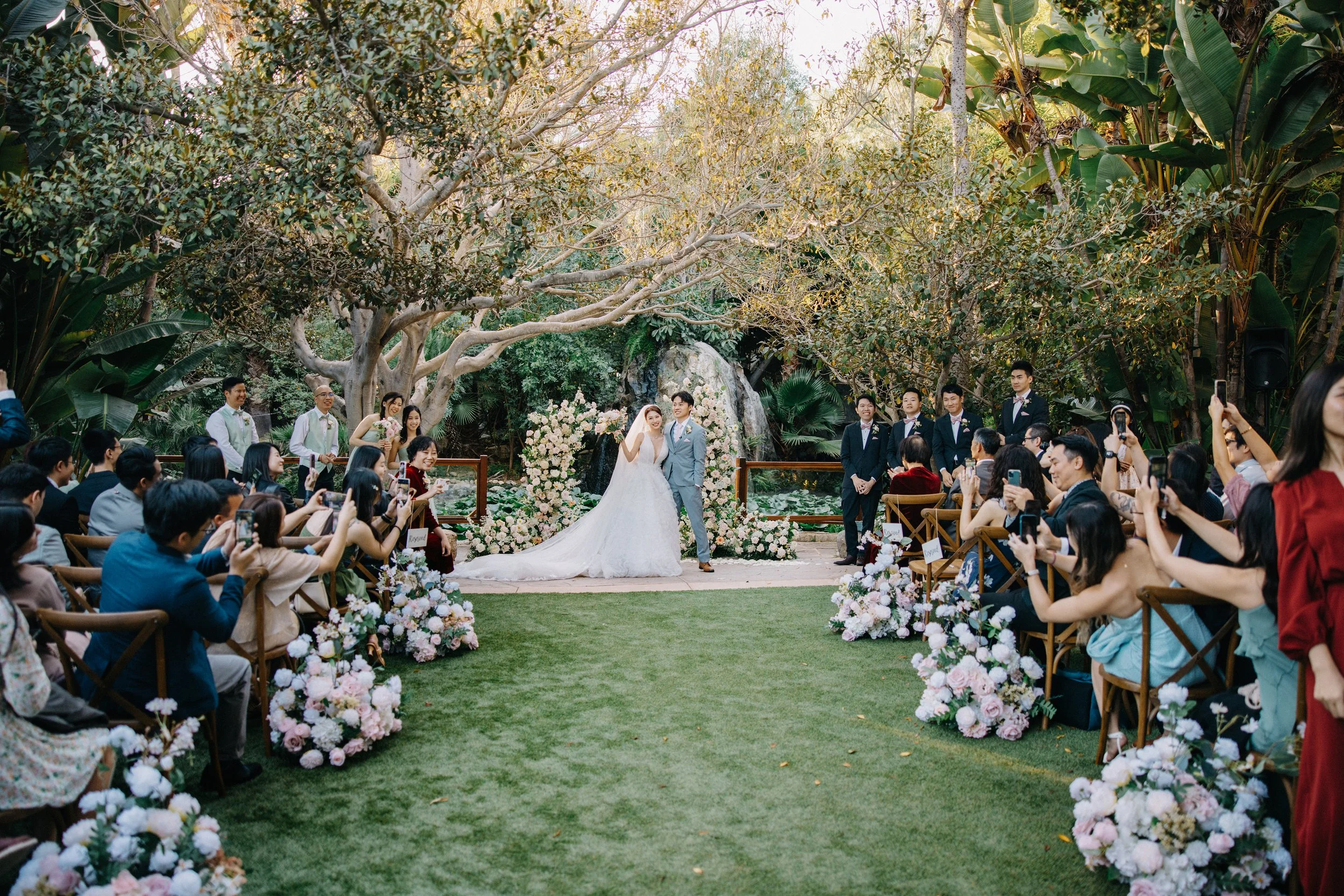 Wedding ceremony outdoors with a bride and groom standing in front of an arch decorated with white flowers, surrounded by lush greenery and trees. Guests seated on both sides hold up phones to take photos, with floral arrangements on chairs and a pon