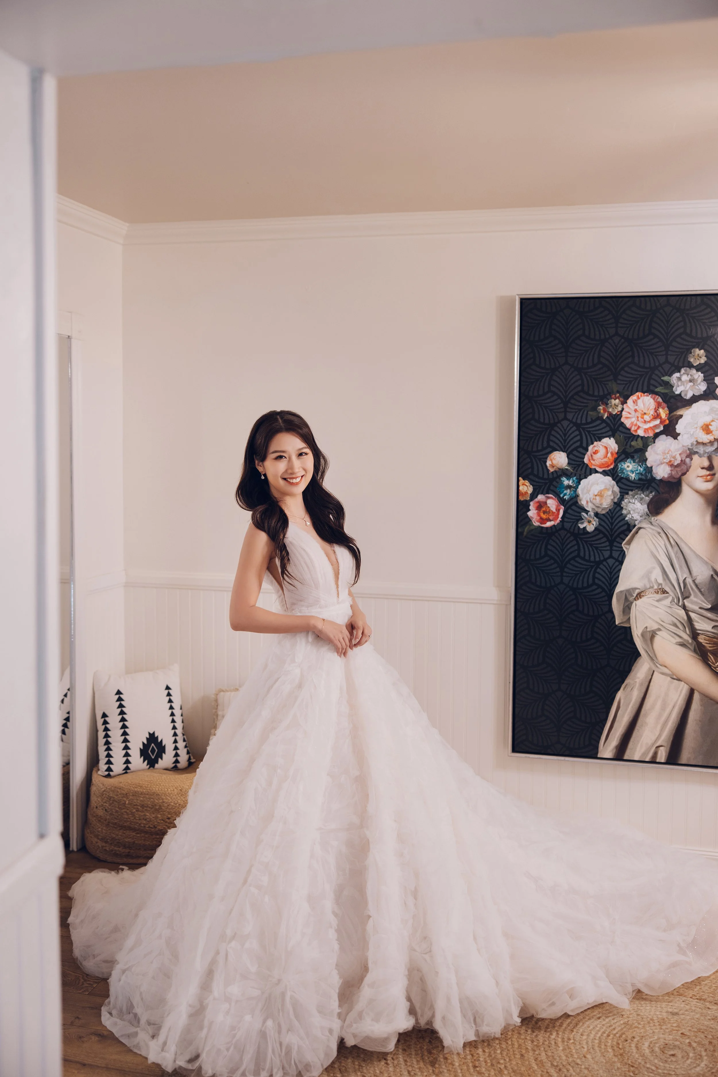 A woman in a white wedding dress smiling in a room with a large portrait on the wall, a mirror reflecting part of the room, and decorative cushions.