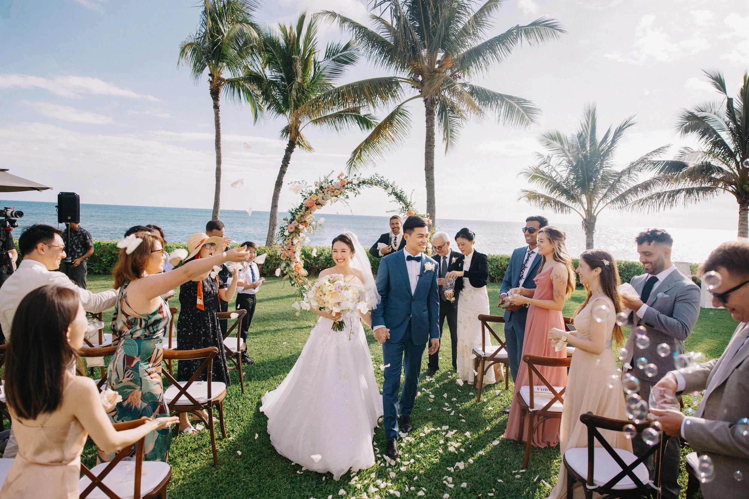 A wedding ceremony outdoors by the ocean with palm trees, where a bride in a white wedding gown and a groom in a blue suit walk hand-in-hand down the aisle, surrounded by guests celebrating with bubbles.