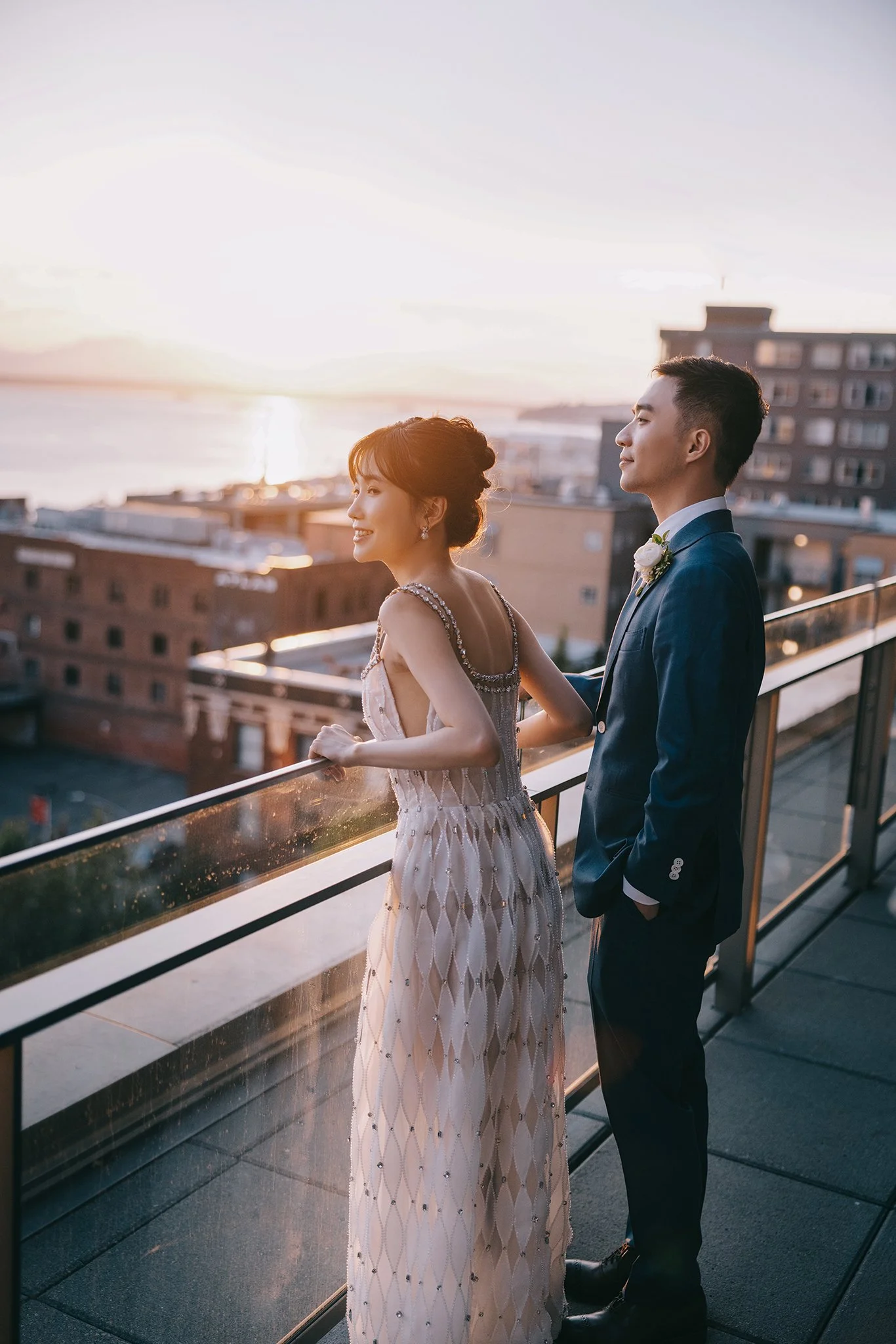 A newlywed couple stands on a rooftop balcony at sunset. The bride, wearing a beaded gown, smiles softly while looking to her right. The groom, dressed in a dark suit with a white shirt and boutonniere, stands beside her with hands in his pockets, lo