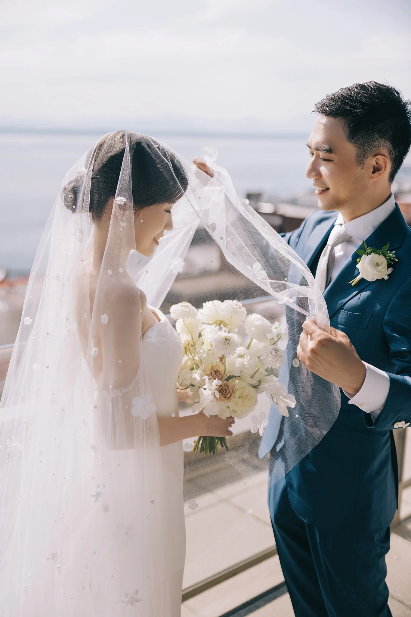 A bride and groom on their wedding day, with the groom lifting the bride's veil outdoors near a body of water, the bride holding a bouquet of white and blush flowers.