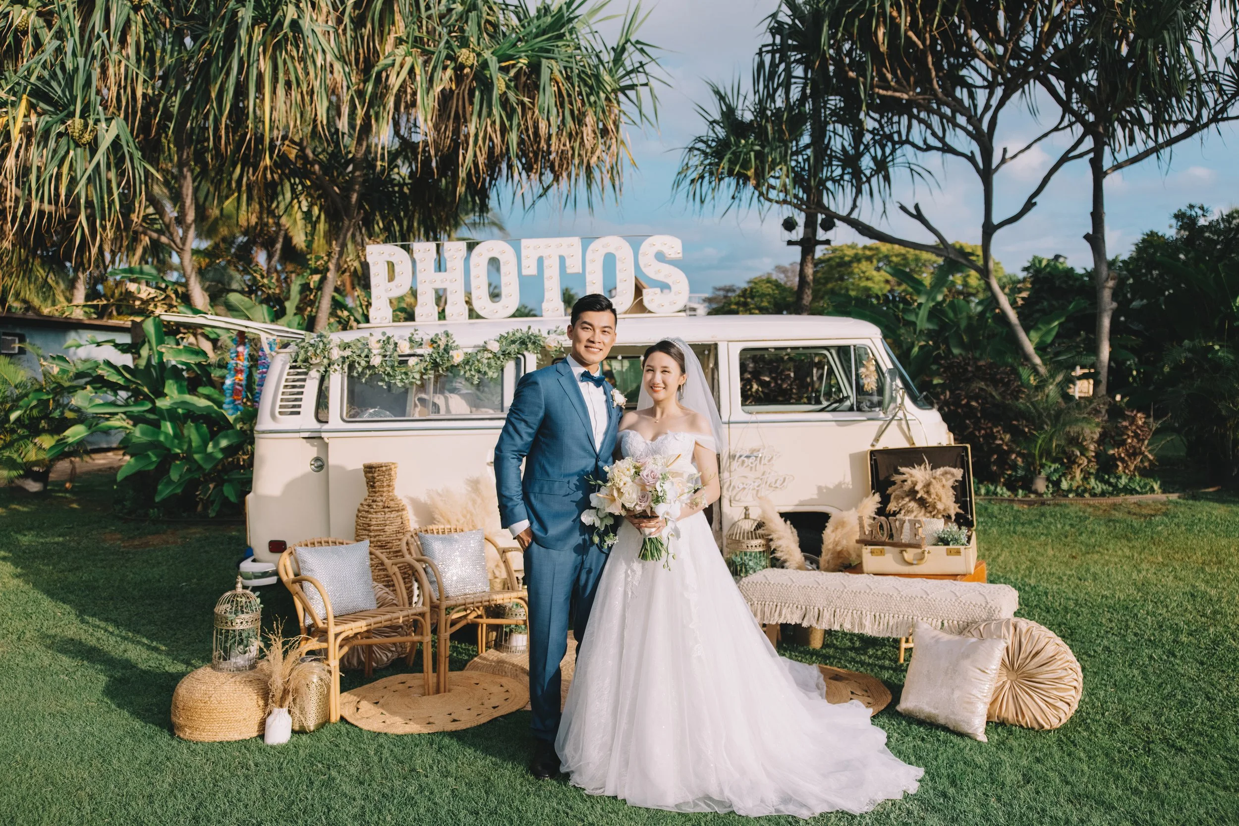 A wedding couple standing outdoors in front of a vintage van with a large illuminated sign that says "PHOTOS" on top. The bride is holding a flower bouquet and the groom is wearing a blue suit. The scene is decorated with rustic and bohemian-style fu