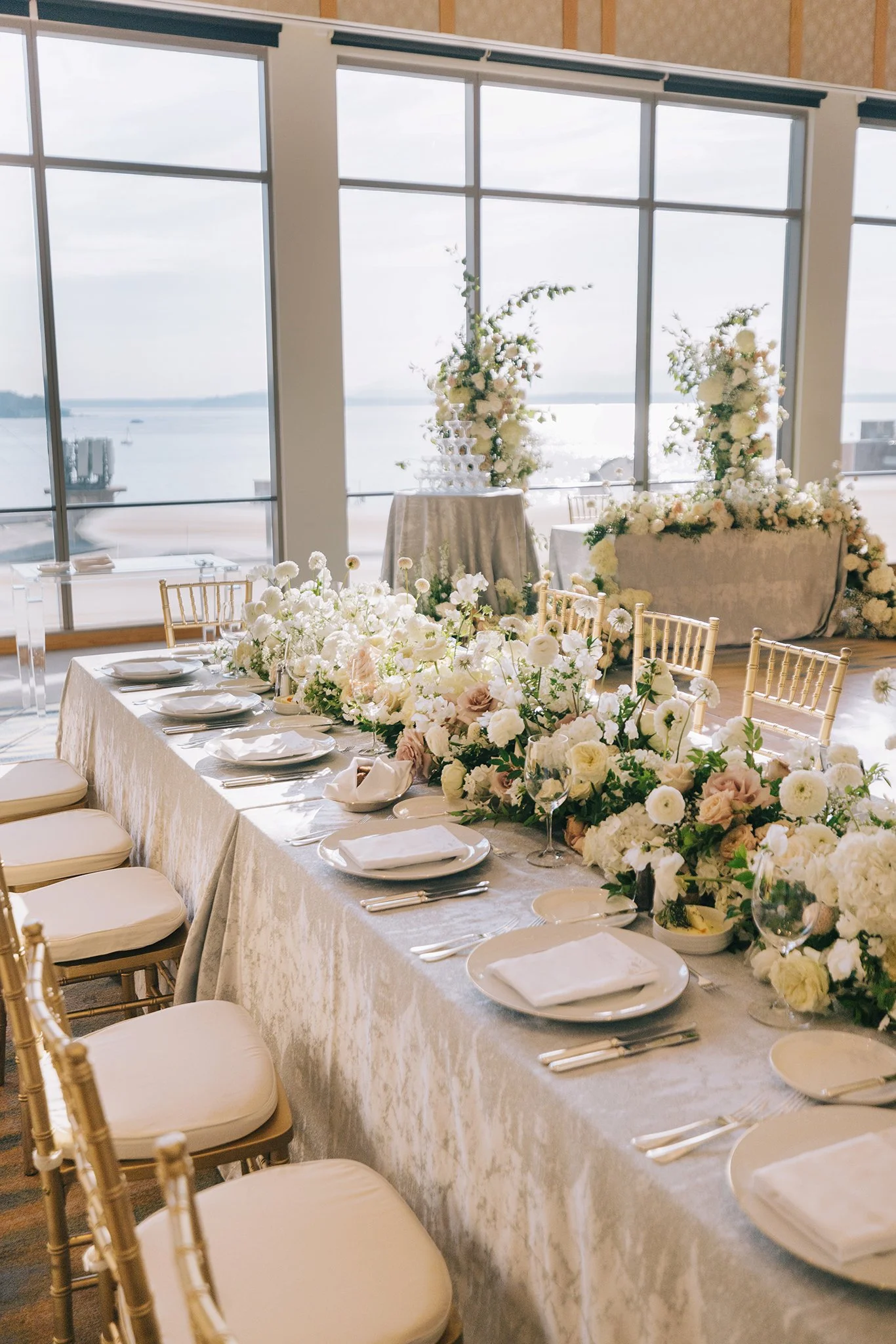 Elegant banquet table decorated with white and blush flowers, set with white plates, silverware, and glassware, overlooking large windows with a waterfront view.