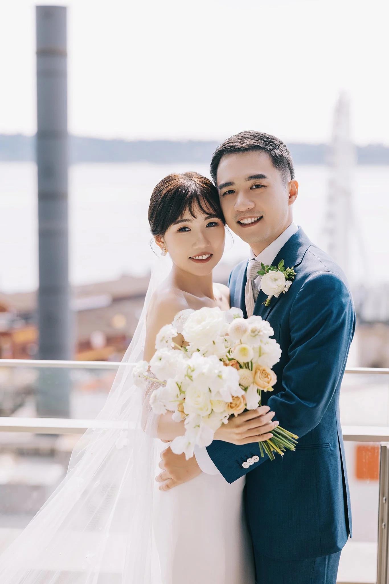 A bride and groom smiling together on their wedding day, outdoors, with water and boats in the background, holding a bouquet of white and blush flowers.