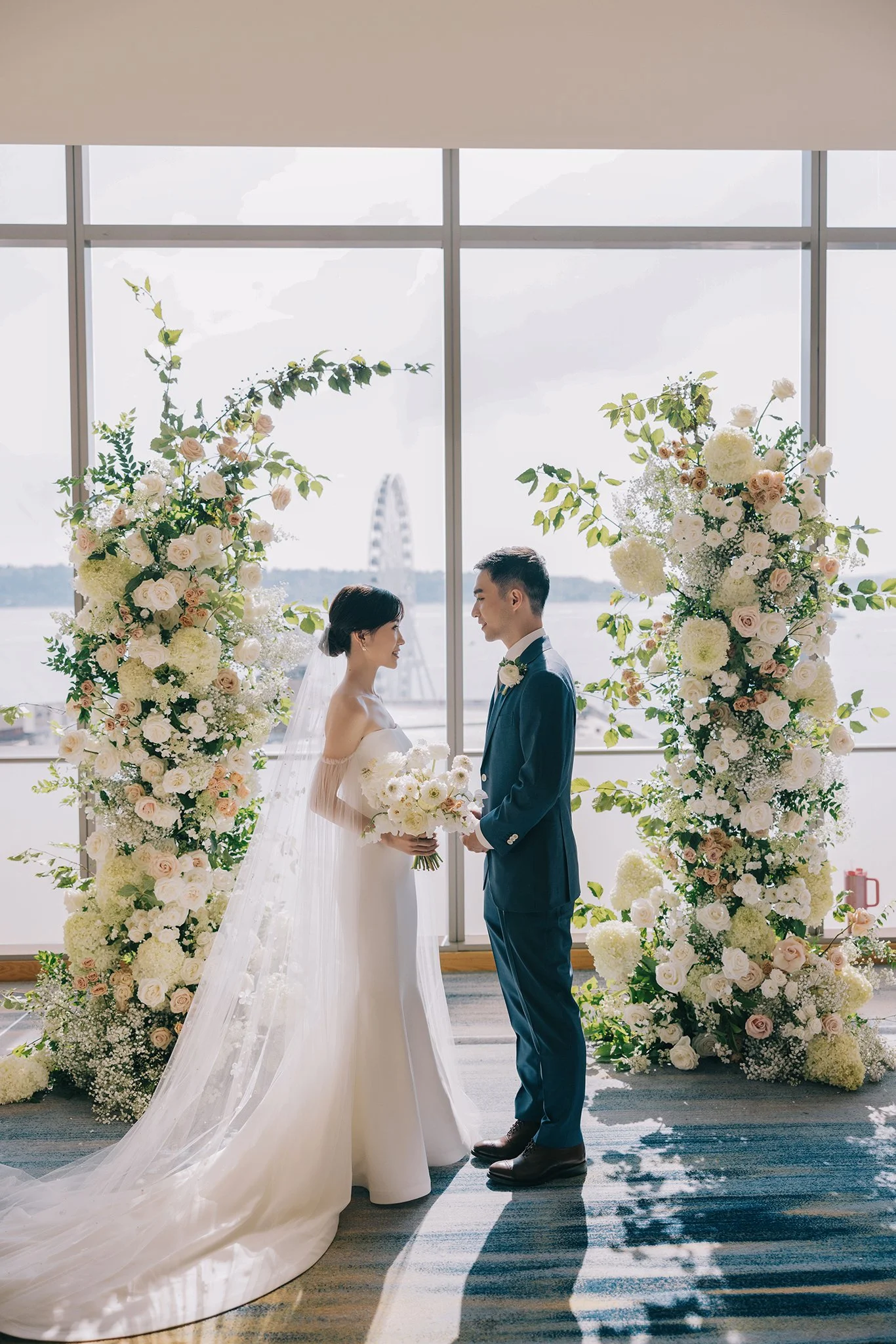 A bride and groom stand facing each other during their wedding ceremony, surrounded by floral arches with white and blush roses, set indoors by large windows with a view of a Ferris wheel outside.