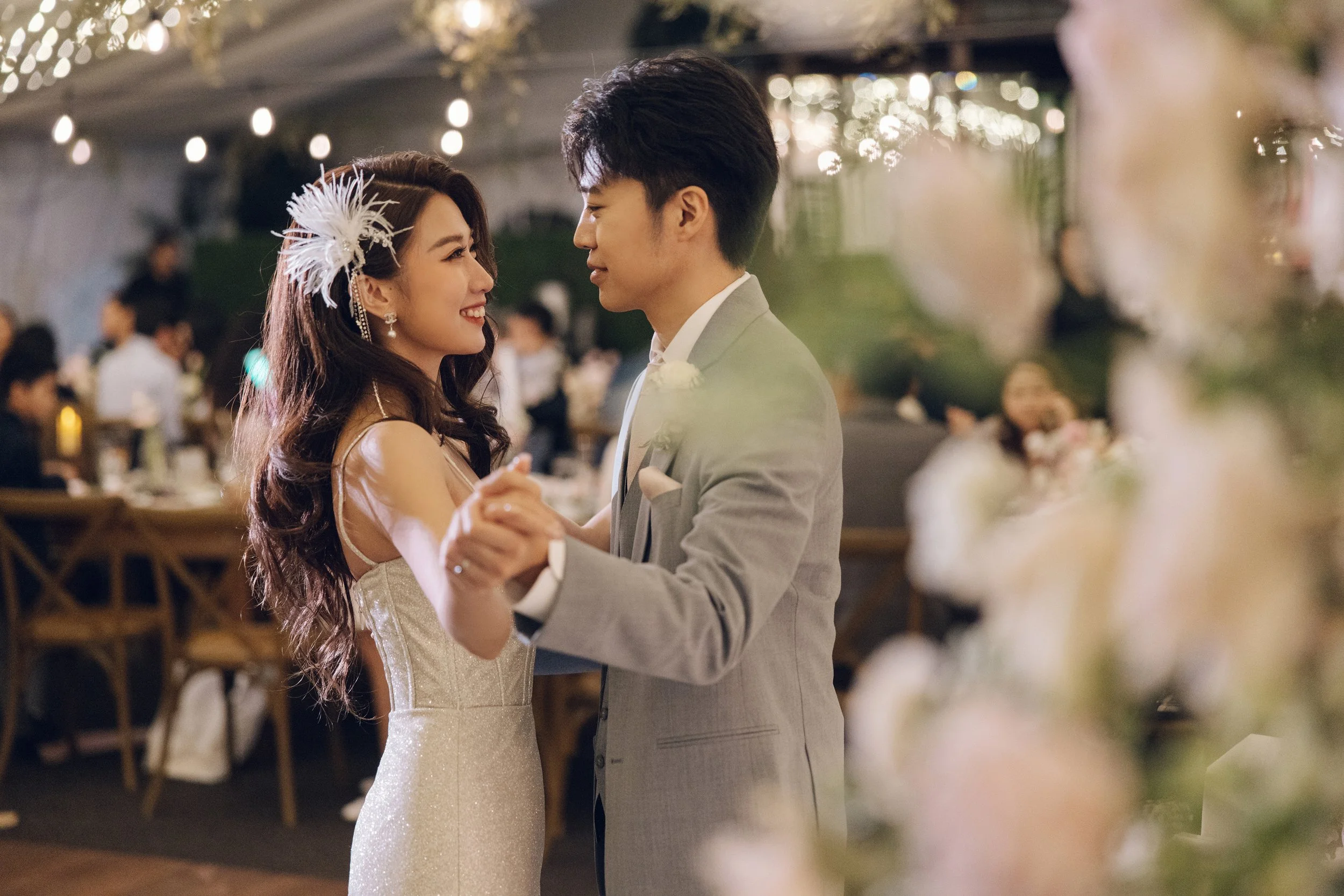 A bride and groom dancing at their wedding reception, smiling at each other, with guests seated at tables in the background.