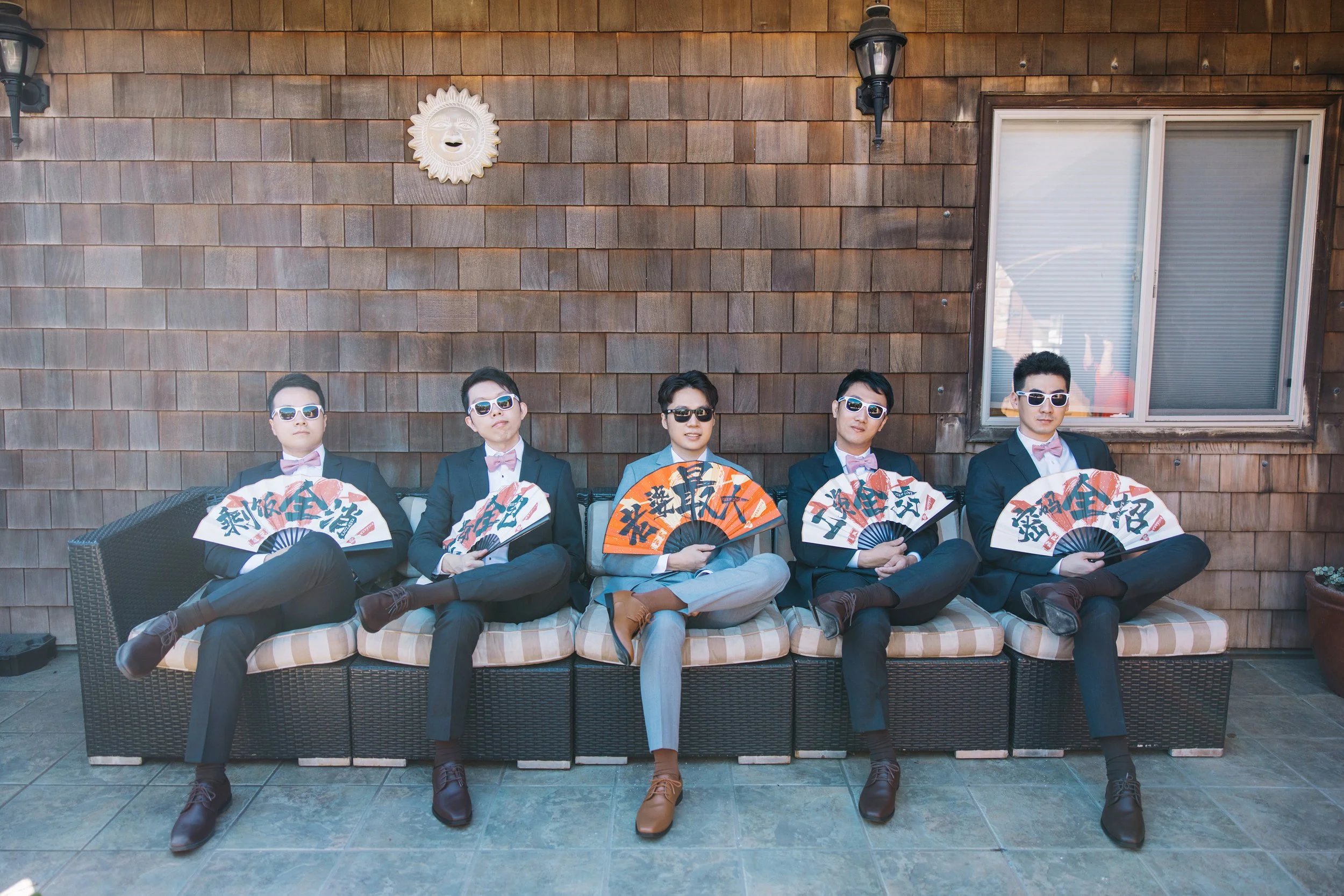 Five young men in suits and sunglasses sitting on a patio sofa, each holding a folding fan with Chinese characters, against a wood-paneled wall with a sun face decoration and a window.