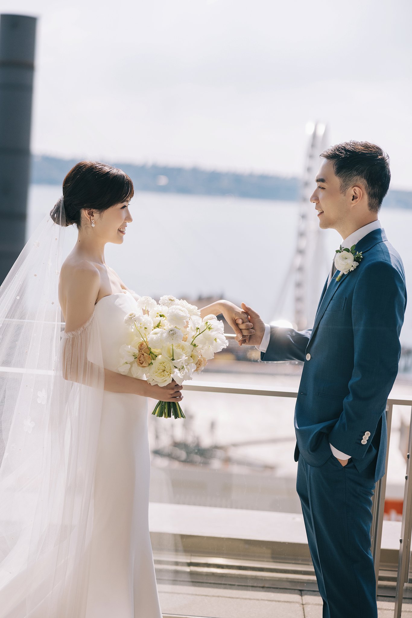 A bride and groom holding hands and smiling at each other outdoors near a body of water during their wedding, with the bride holding a bouquet of white and beige flowers.