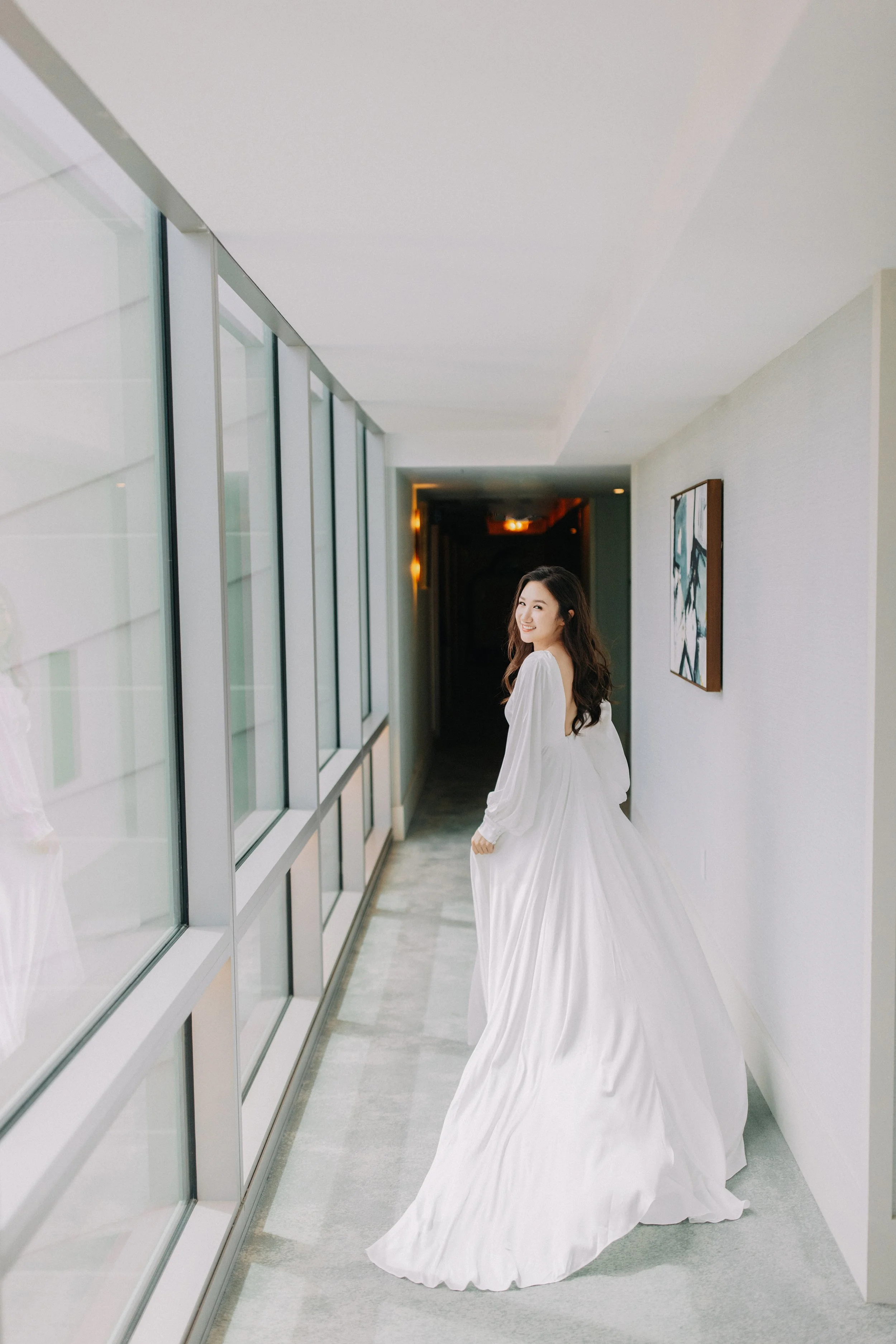 A smiling woman wearing a white wedding dress walking down a brightly lit corridor with large windows on her left and abstract artwork on her right wall.