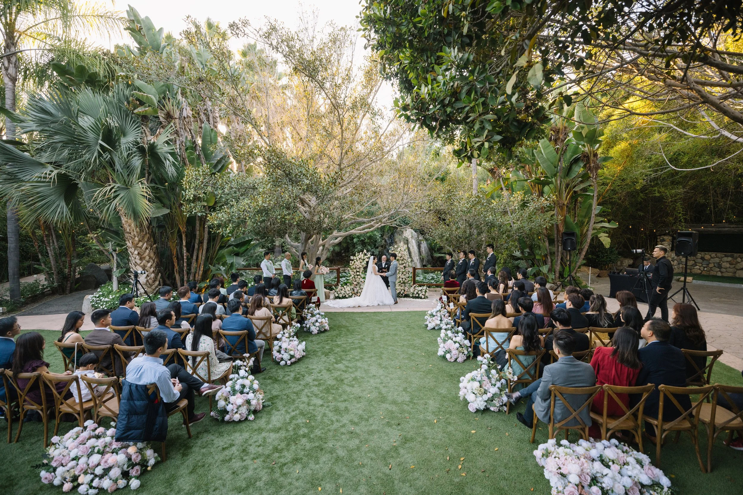 Outdoor wedding ceremony with bride and groom exchanging vows at altar surrounded by floral arrangements and guests seated on both sides, lush greenery and large trees in the background.