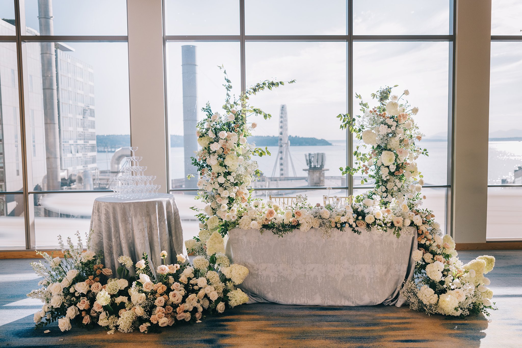 Elegant floral wedding display with white and blush roses, hydrangeas, and greenery, set against large windows overlooking water and city skyline, with a champagne tower on a round table to the left.