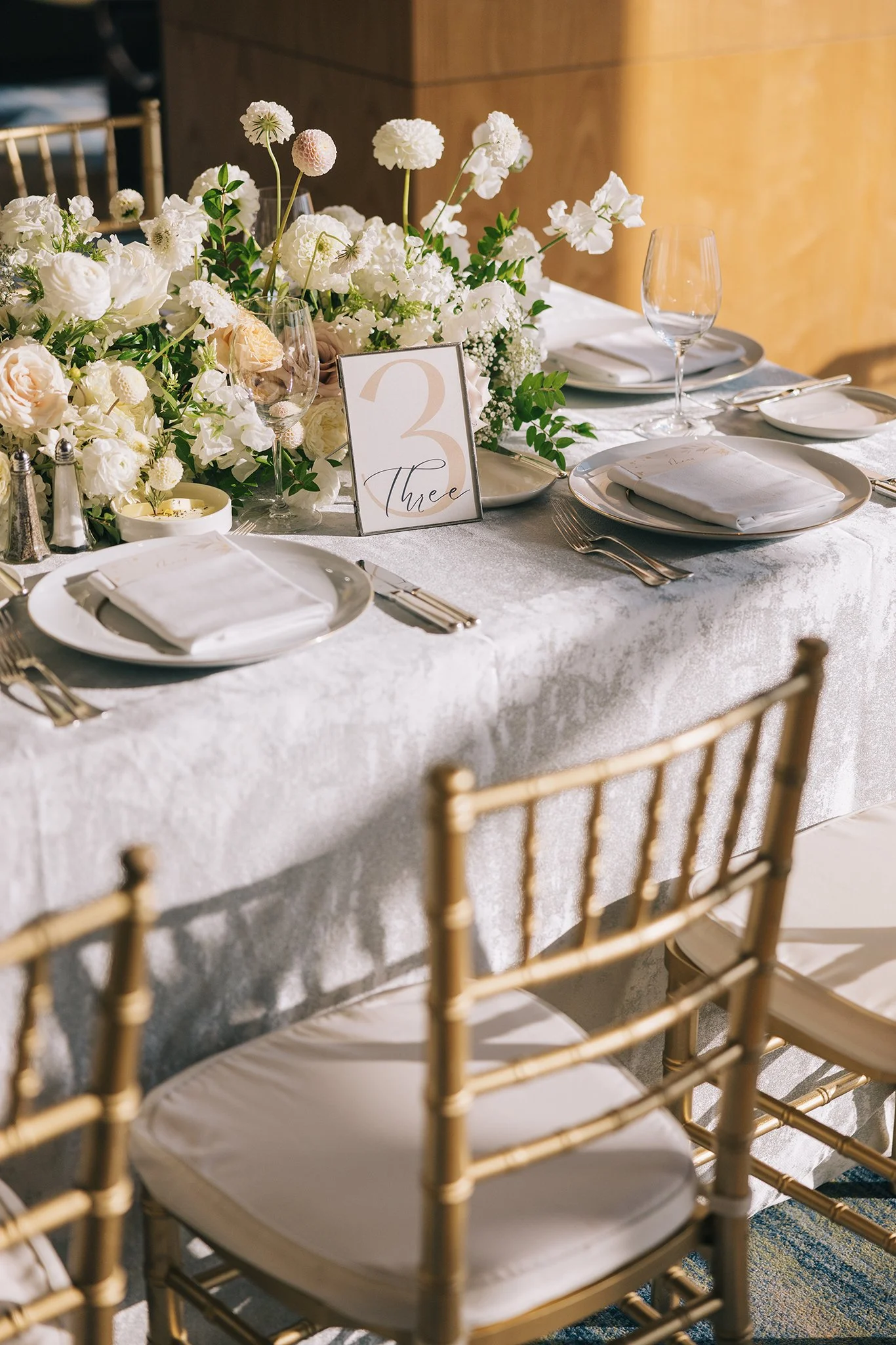 Elegant dining table decorated with white and blush flowers, set with white plates, napkins, silverware, glassware, and a table number card that reads '3' at a formal event.