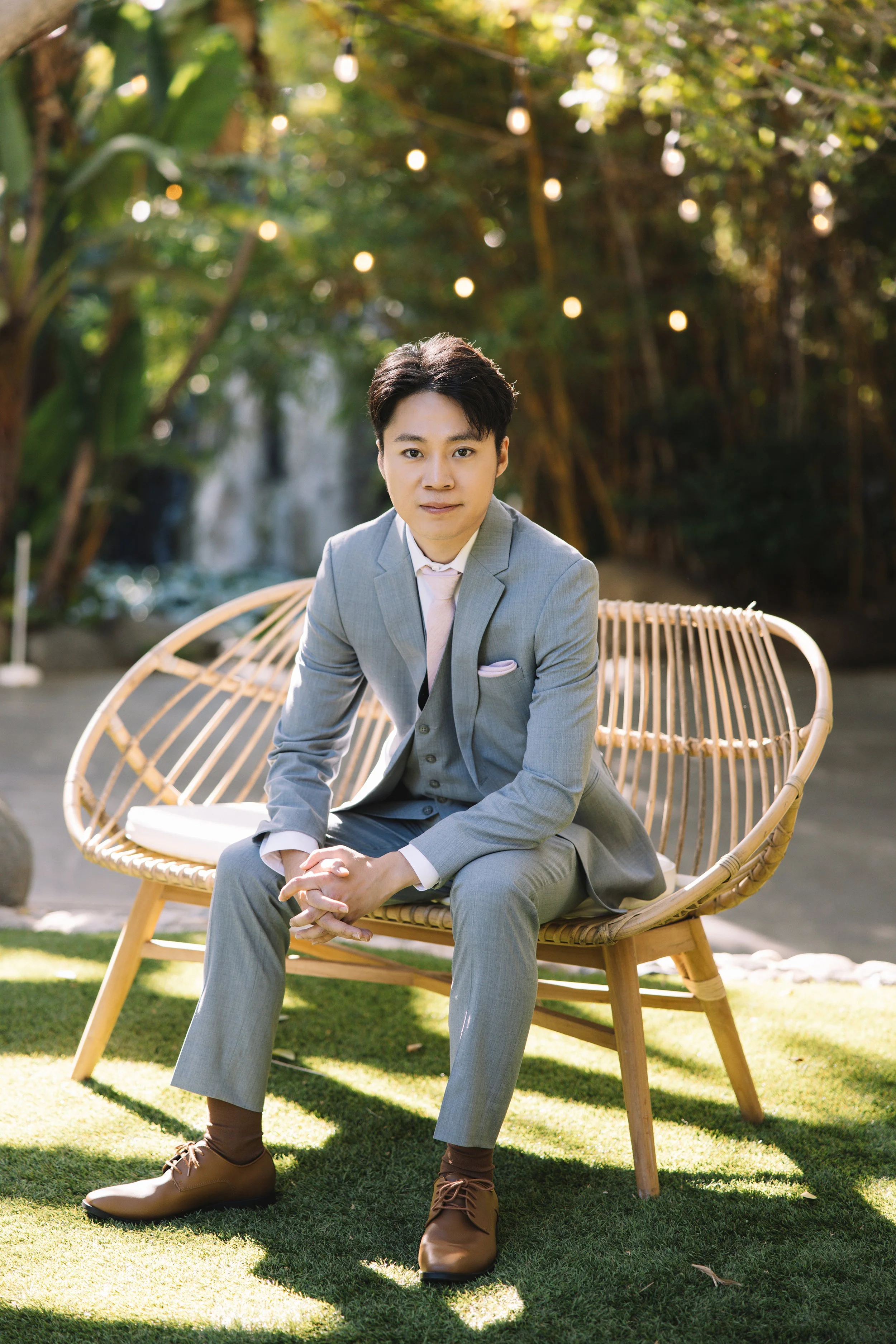 A young man in a gray suit sits on a rattan bench outdoors, with string lights and greenery in the background.
