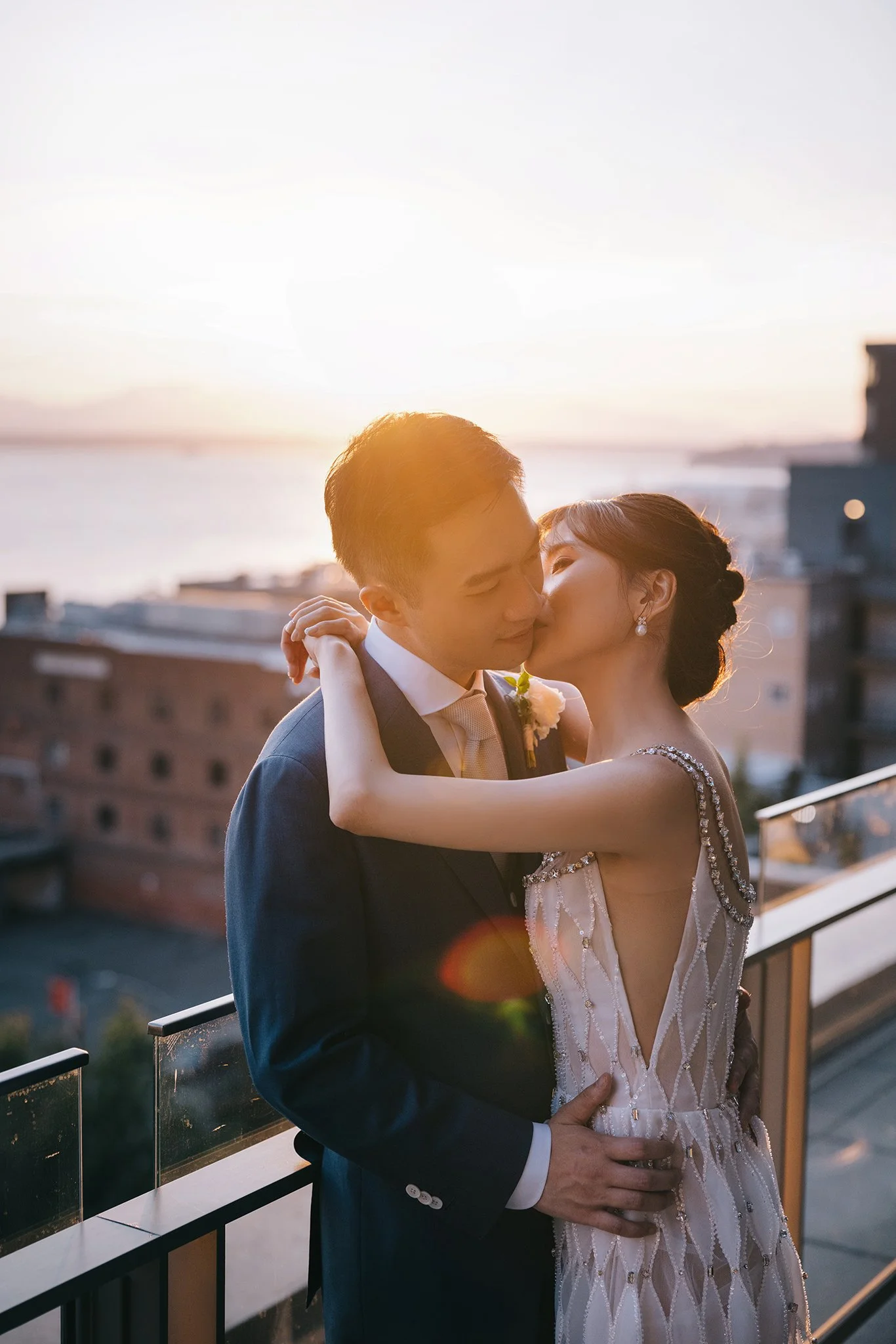 A bride and groom sharing a kiss on a balcony at sunset, with city buildings and water in the background.