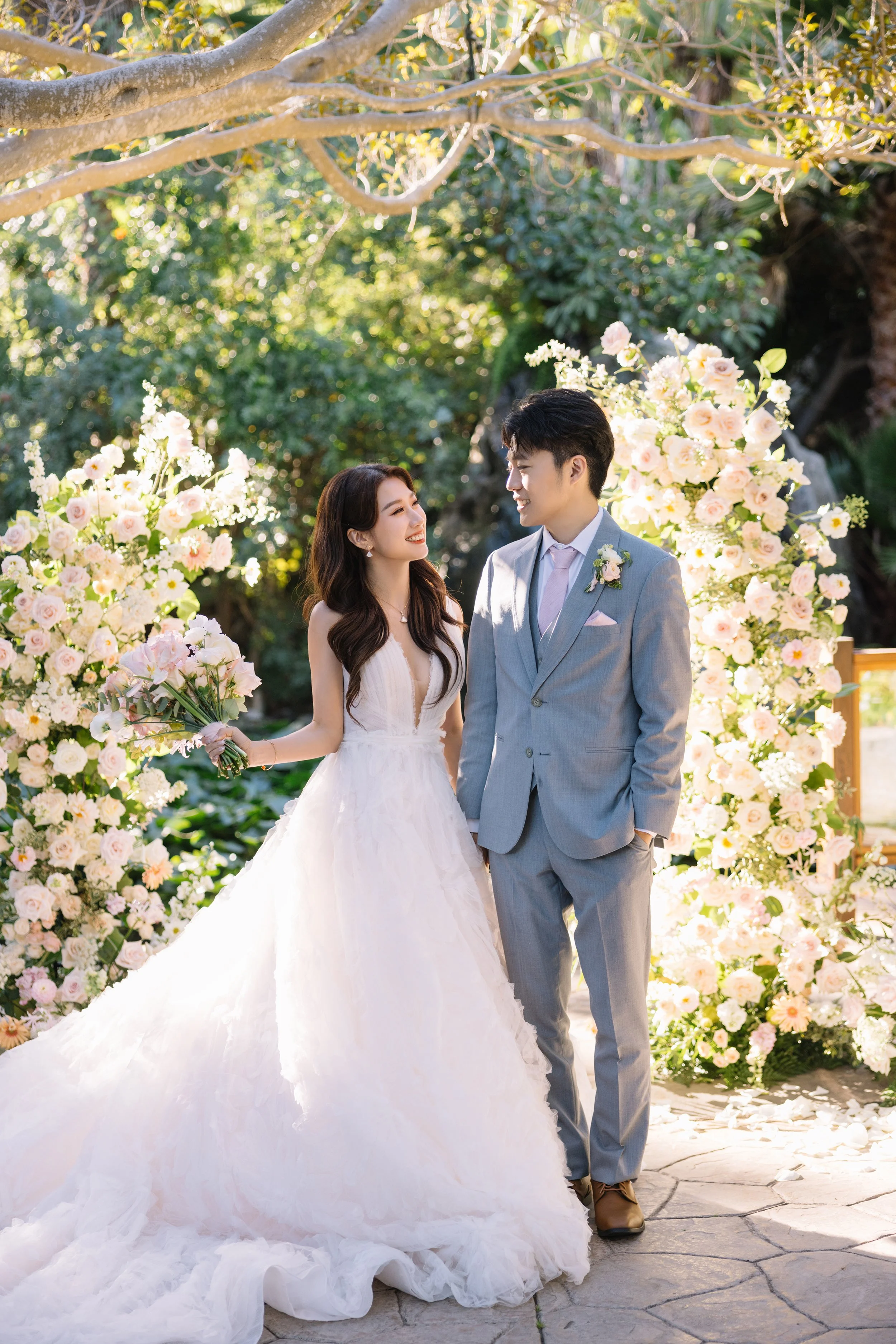 A bride and groom standing together outdoors during their wedding, surrounded by flower arrangements with pink and white flowers, under a large tree with sunlight filtering through.
