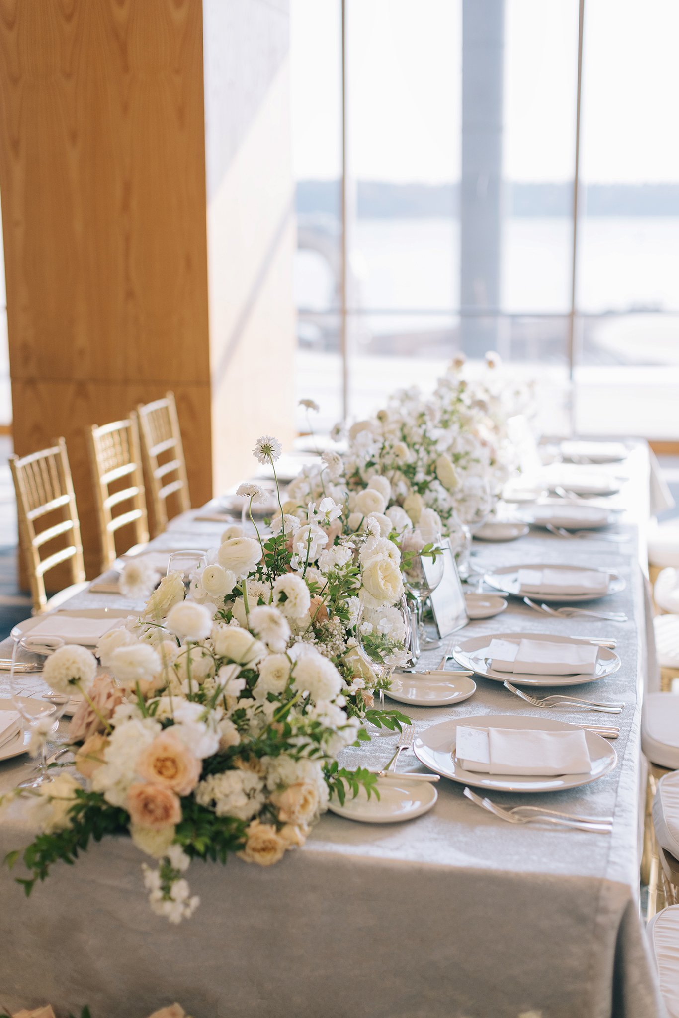 Elegant dining table decorated with white floral centerpiece and neatly arranged plates and cutlery in a bright room with large windows.