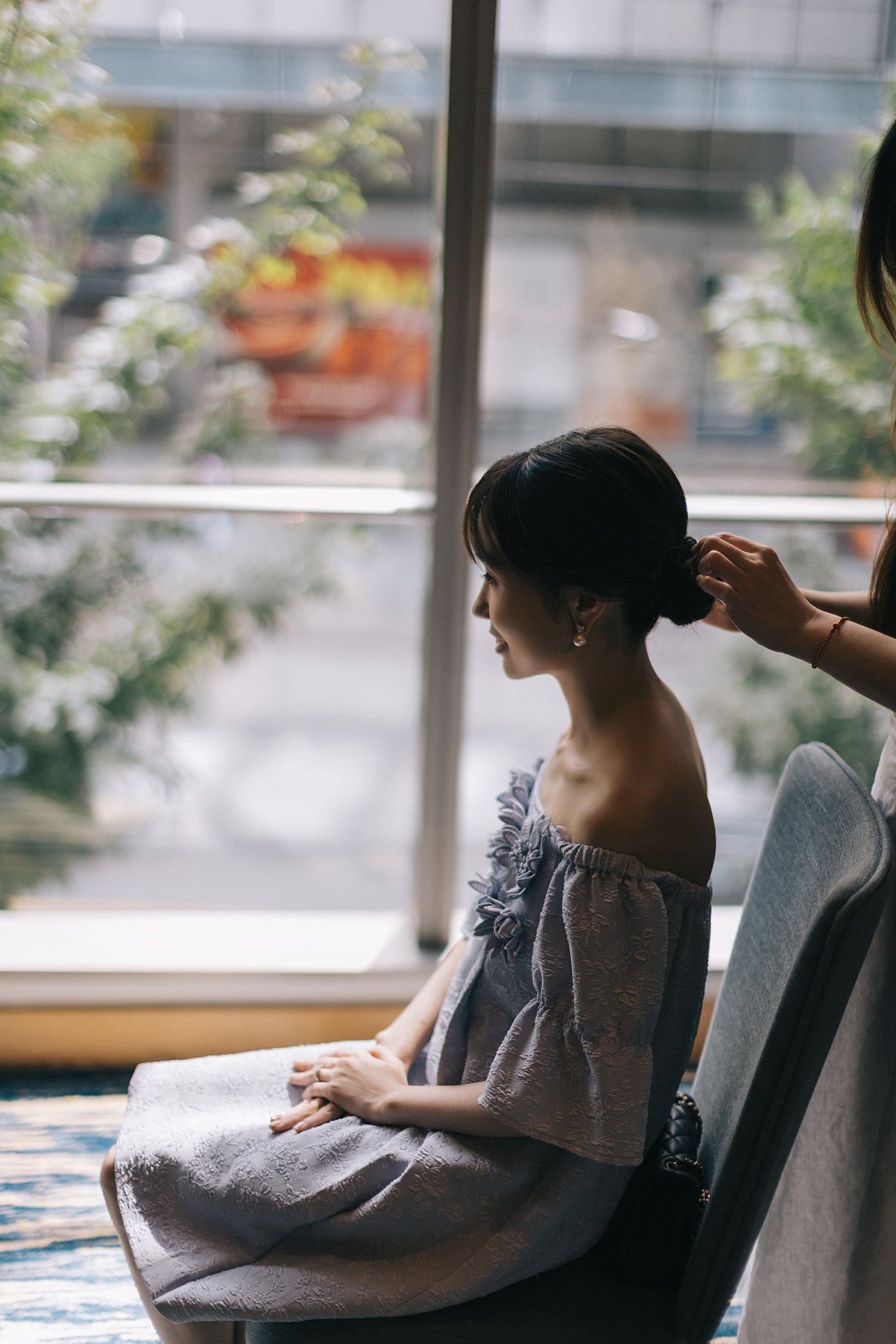 A woman is sitting in a chair by a window, getting her hair styled by a person standing behind her. The woman is wearing an off-the-shoulder, textured dress with floral details.