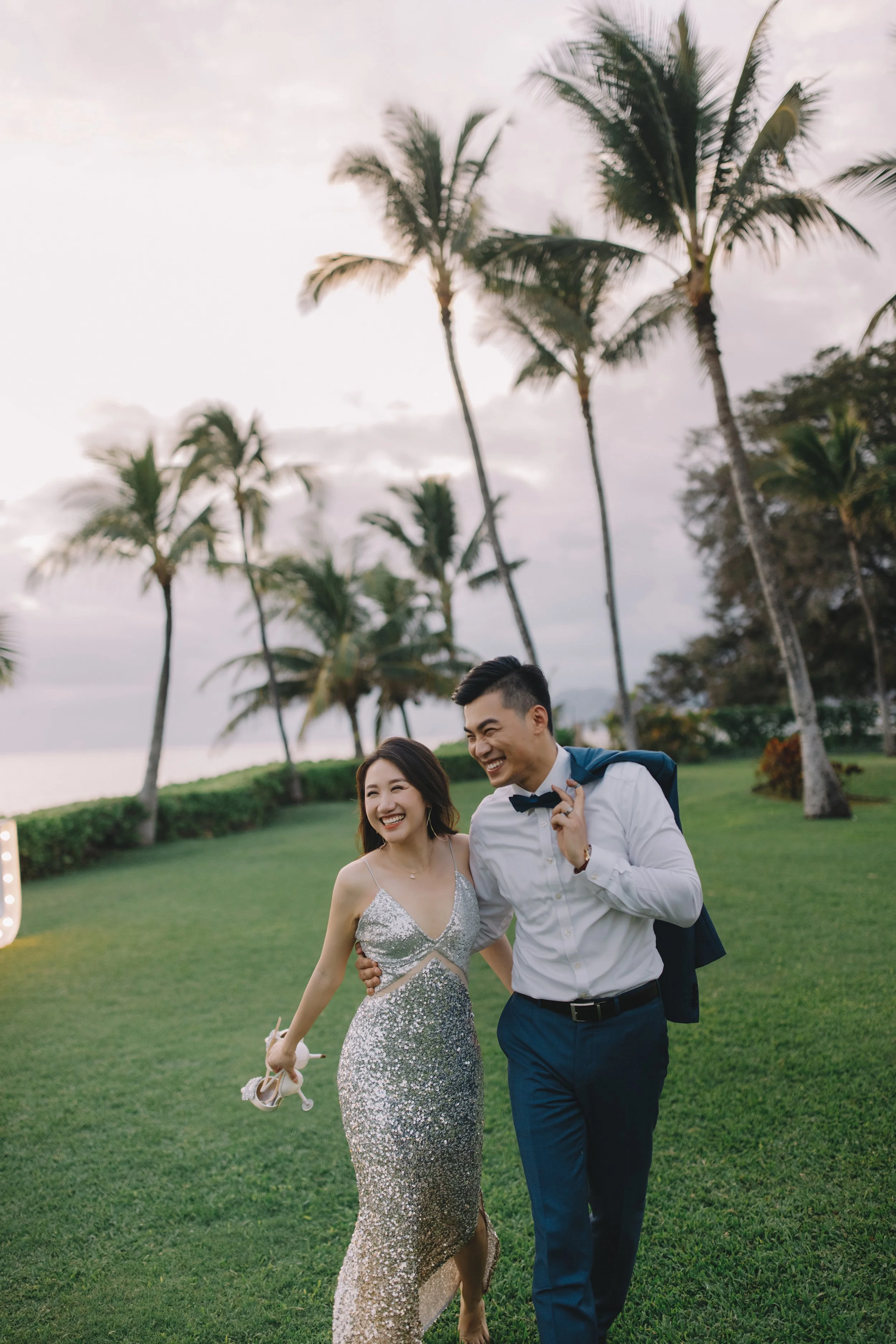 A smiling couple, dressed in formal attire, walking on a grassy area with palm trees in the background, during sunset.
