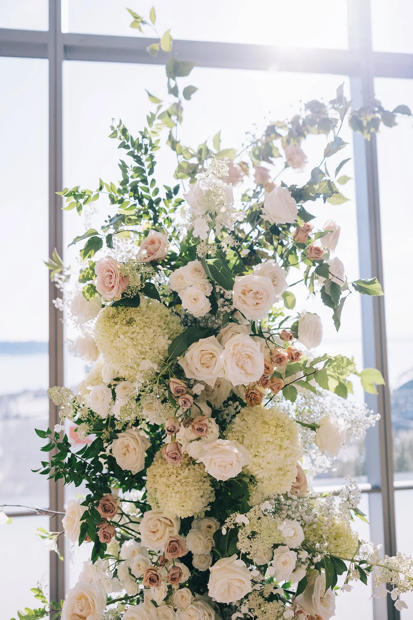 A tall floral arrangement of white and blush roses, hydrangeas, and greenery against a large window background with sunlight.