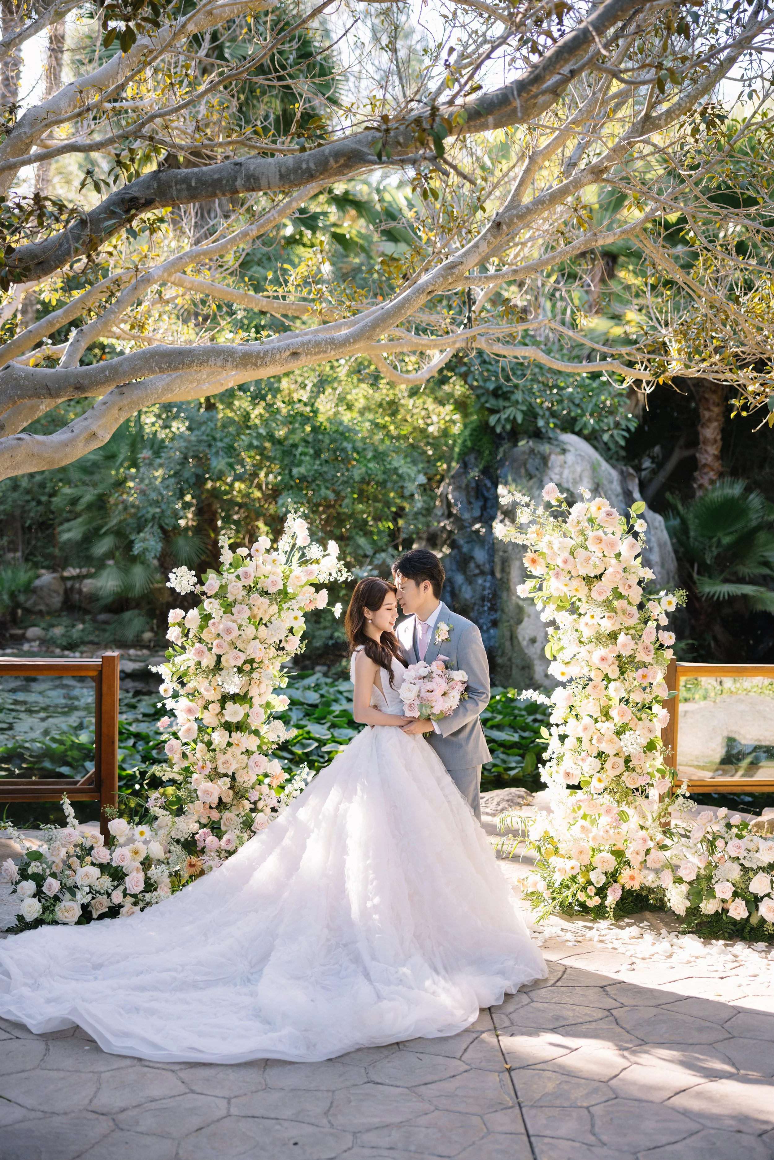 A bride and groom in wedding attire standing close together outdoors, surrounded by pink and white floral arrangements, with trees and rocks in the background.