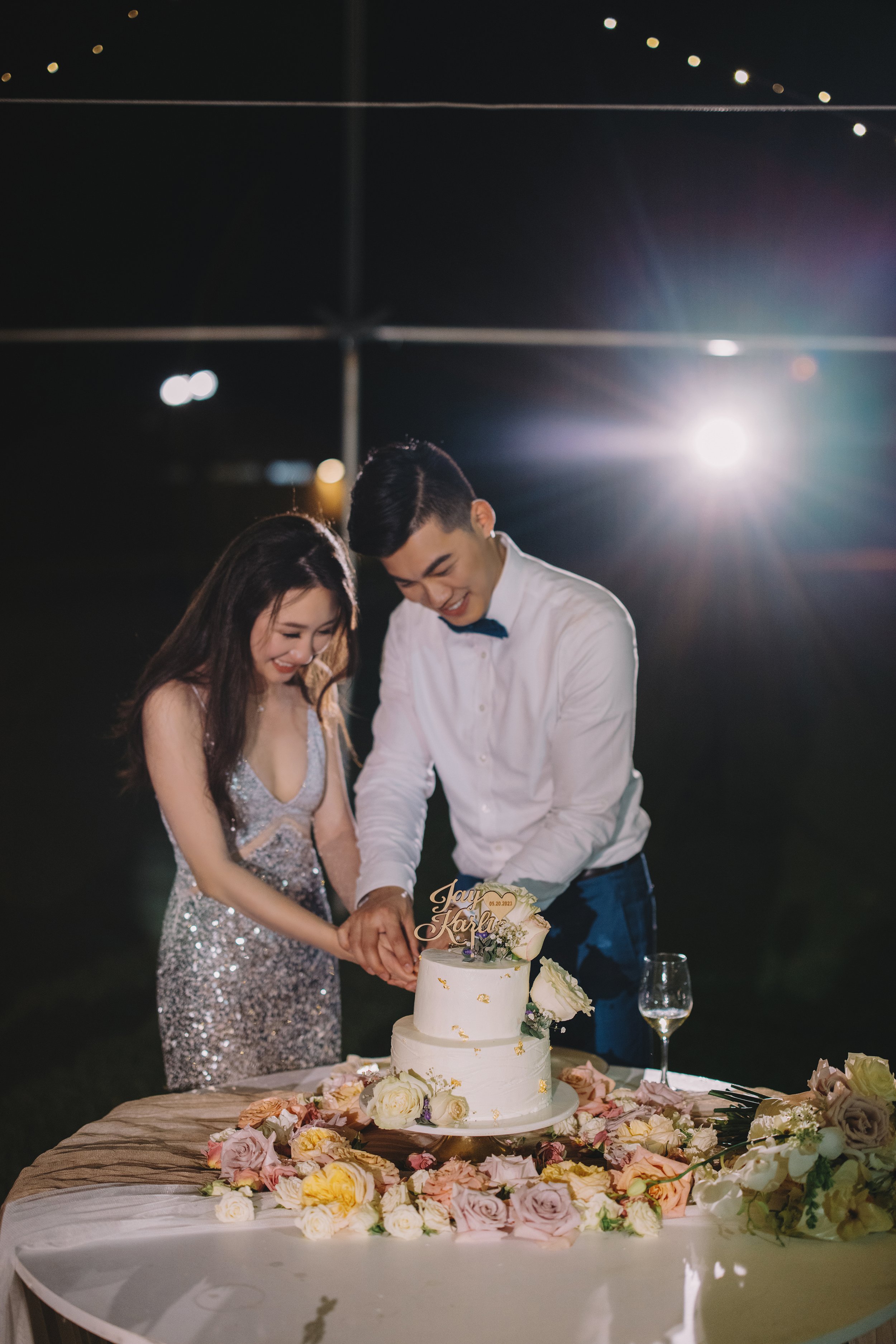 A couple in formal attire cut a wedding cake together at a nighttime celebration, surrounded by flowers and lit by a bright light in the background.