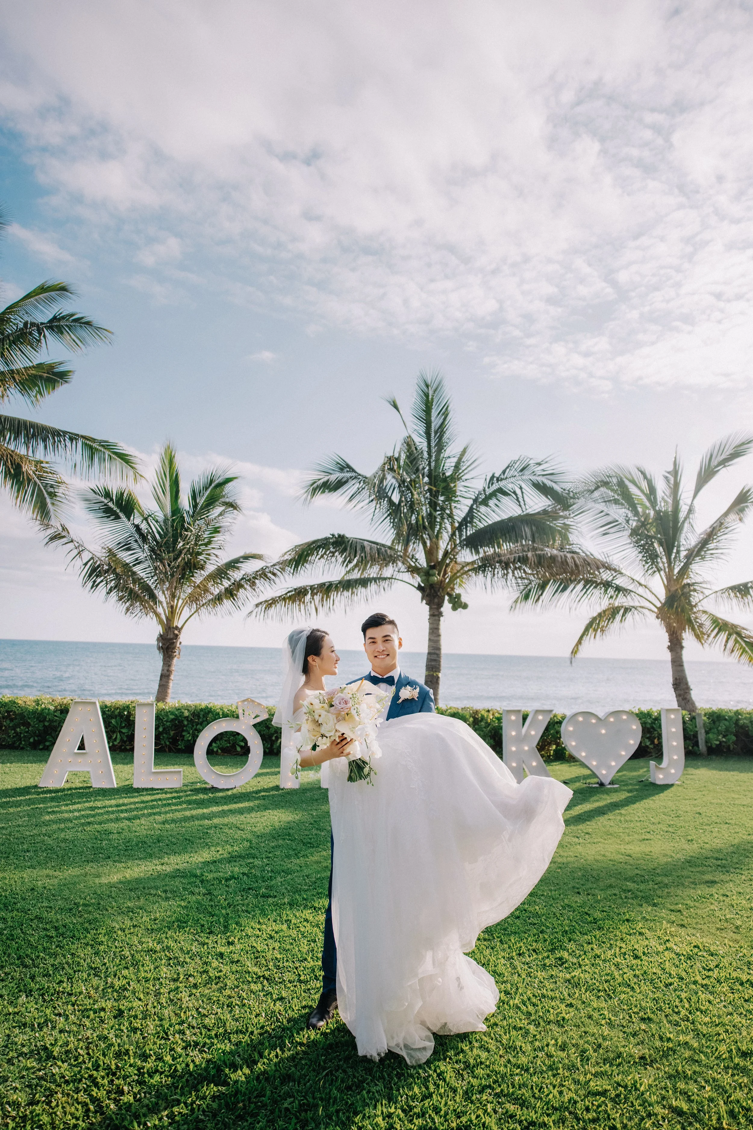 A newlywed couple standing on a grassy area near the ocean, with palm trees in the background and large illuminated letters spelling 'ALOHA' with a heart symbol in the middle.