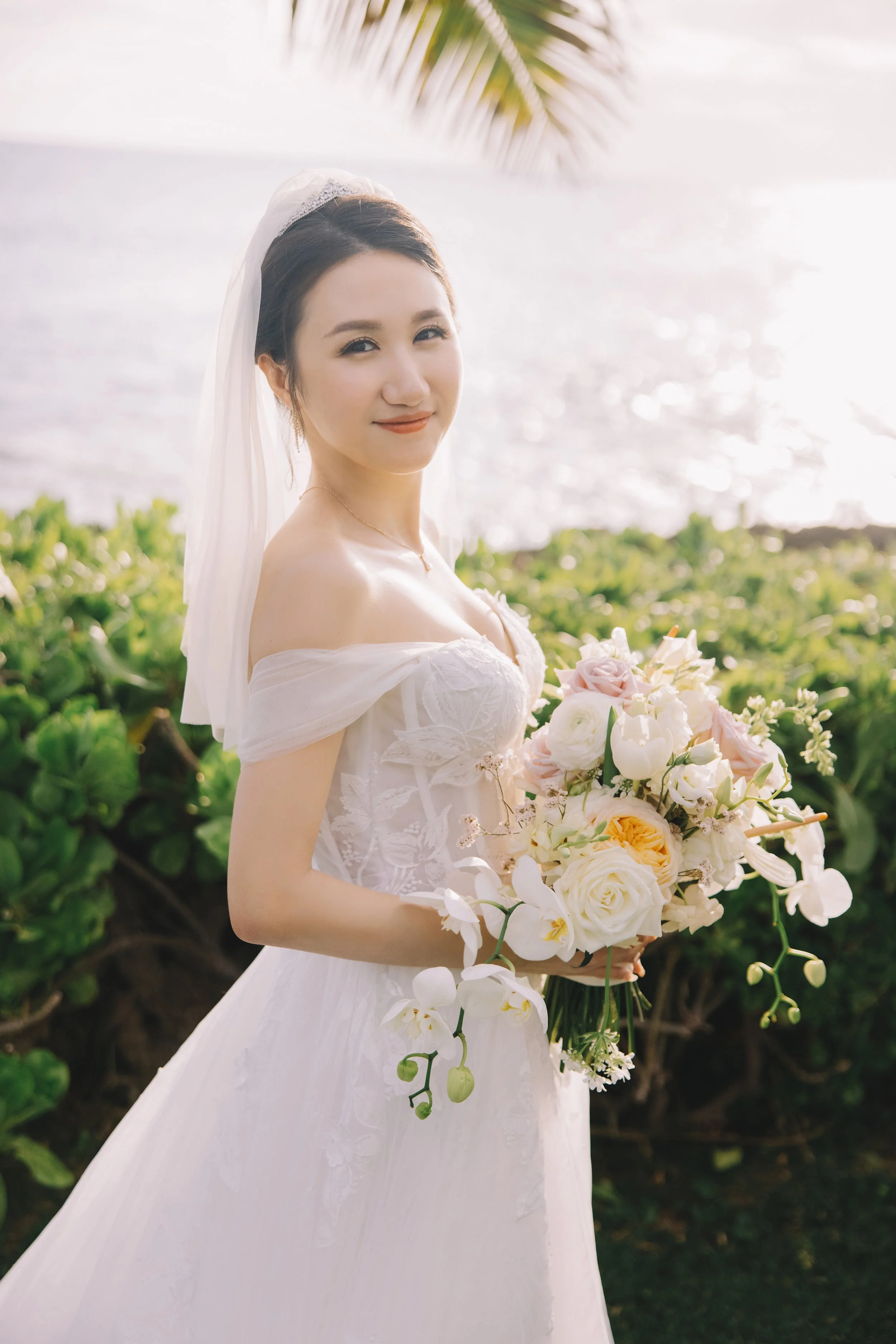 A bride in a white wedding dress holding a bouquet of flowers outdoors near water, with greenery and sunlight in the background.