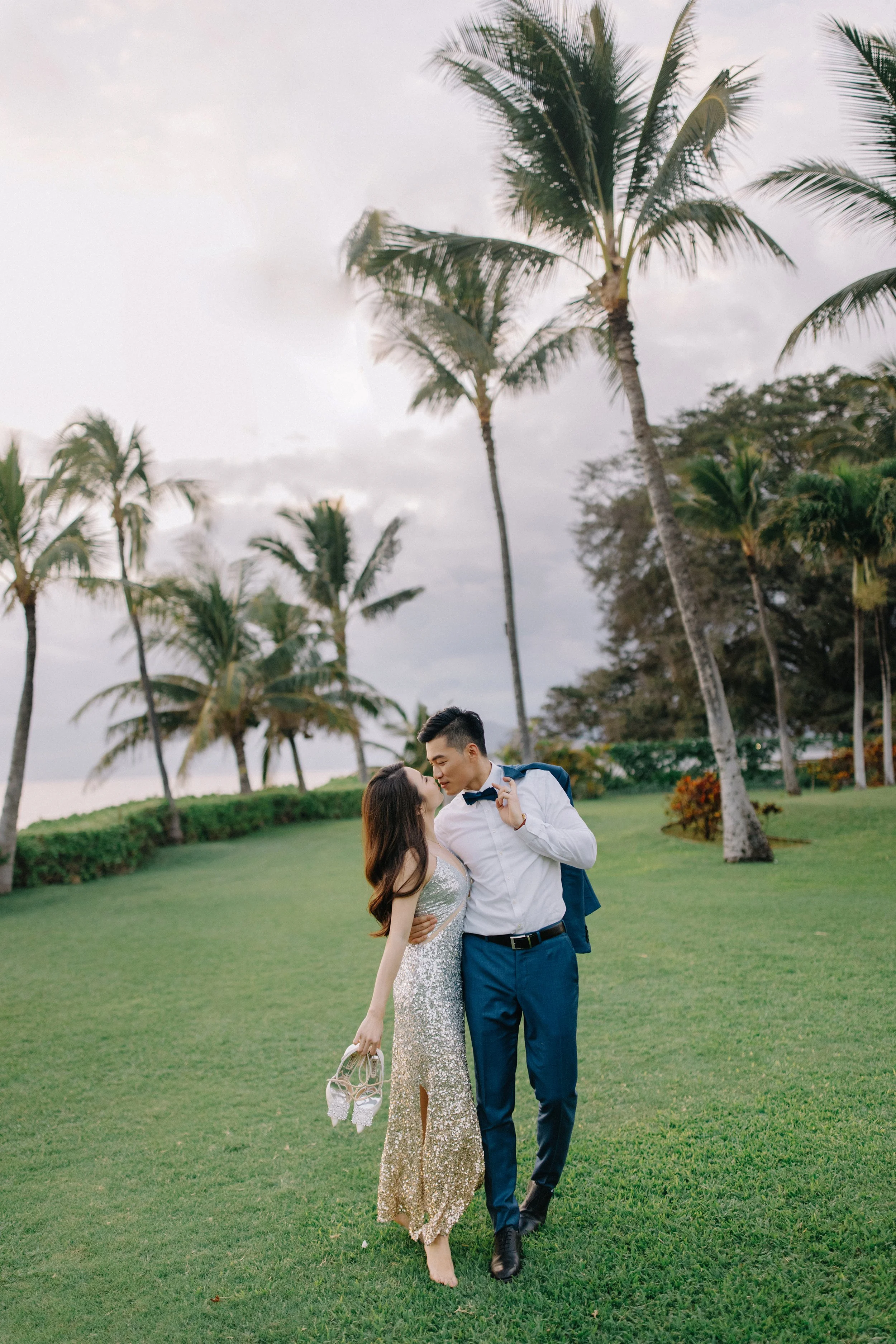 A couple dressed in formal attire sharing a romantic moment on a green lawn with palm trees and a cloudy sky in the background.
