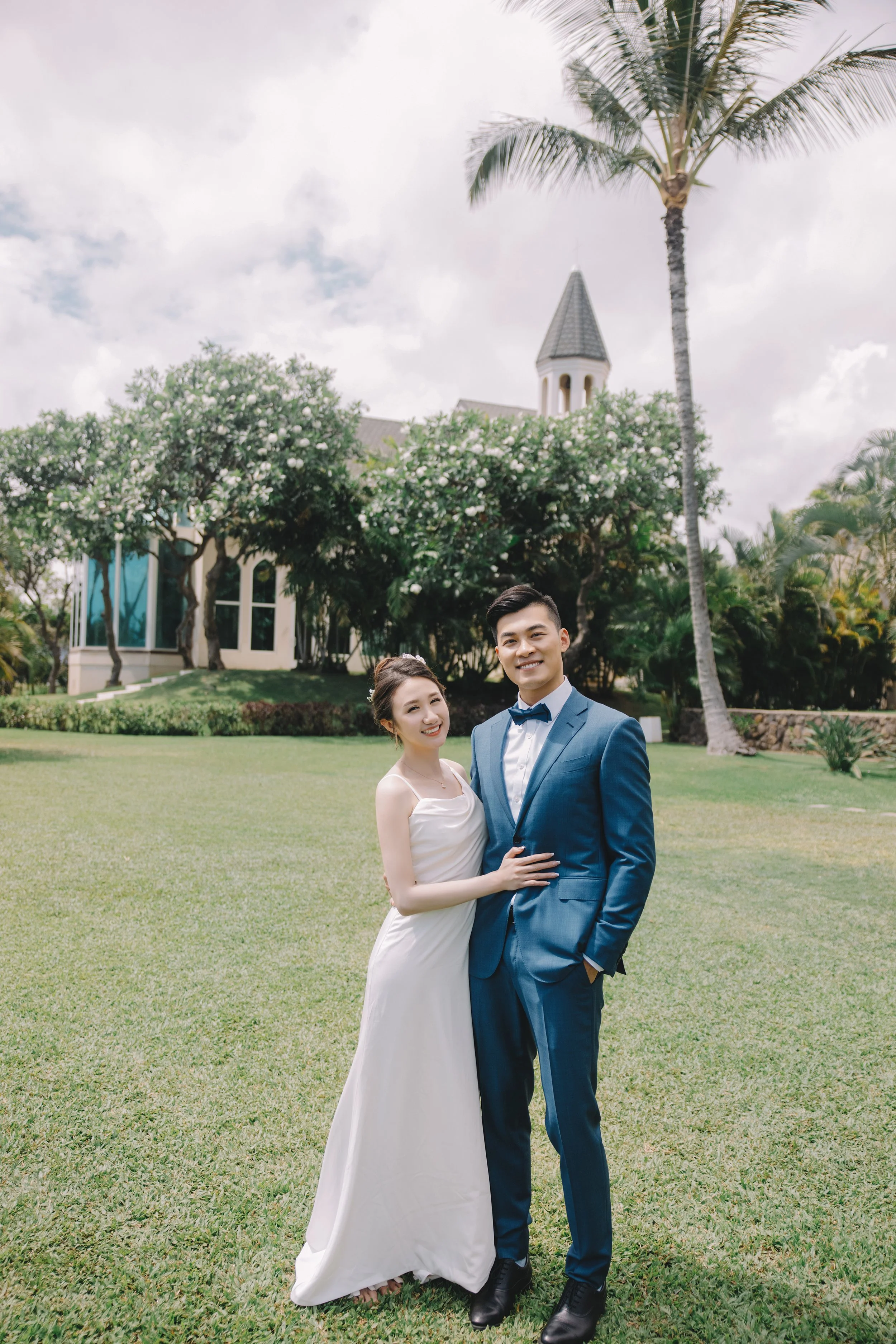 A bride in a white wedding dress and a groom in a blue suit pose together on a lush green lawn with trees, bushes, and a church in the background under a partly cloudy sky.