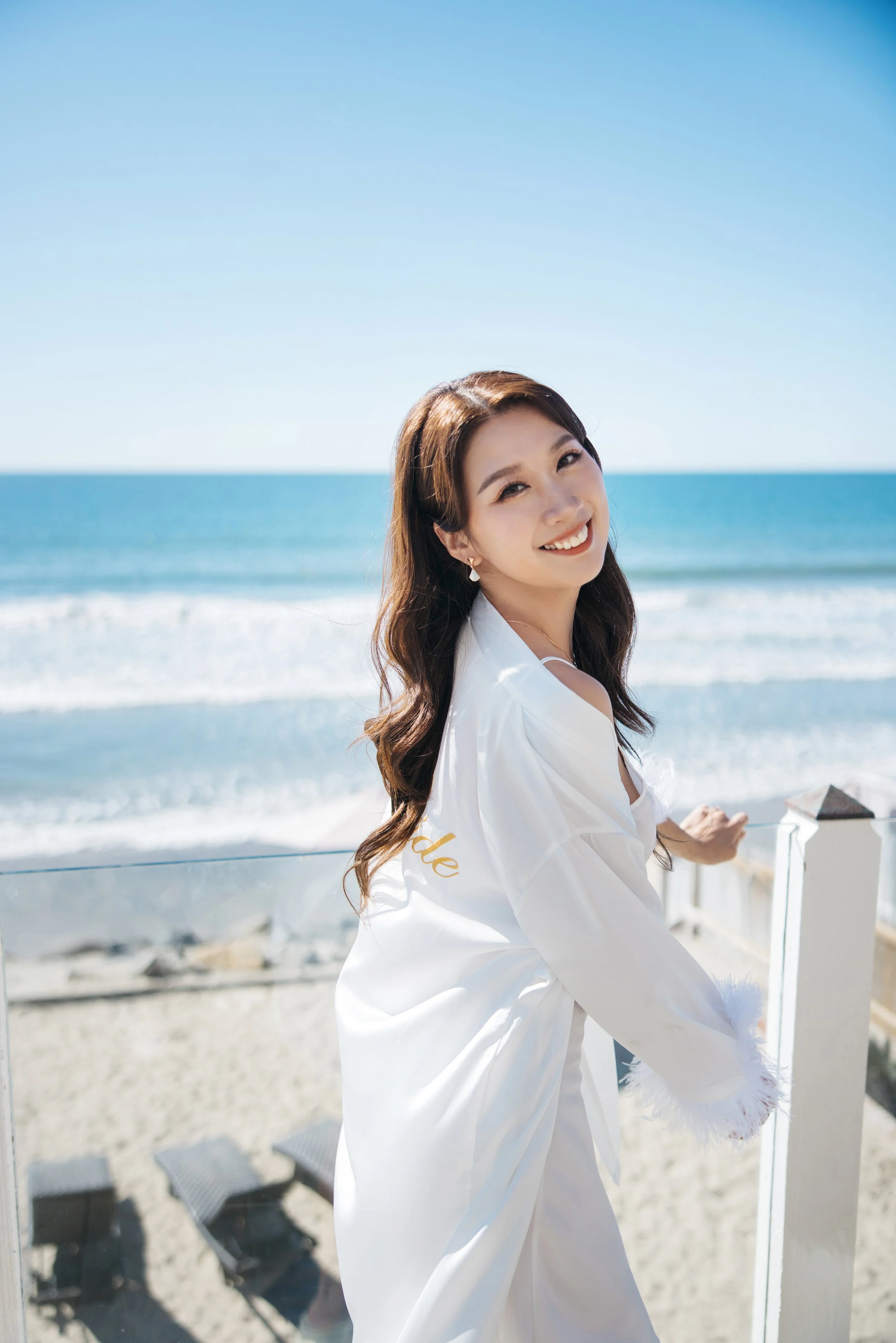 A young woman with long brown hair, smiling, wearing a white robe, standing on a balcony overlooking the ocean on a sunny day.