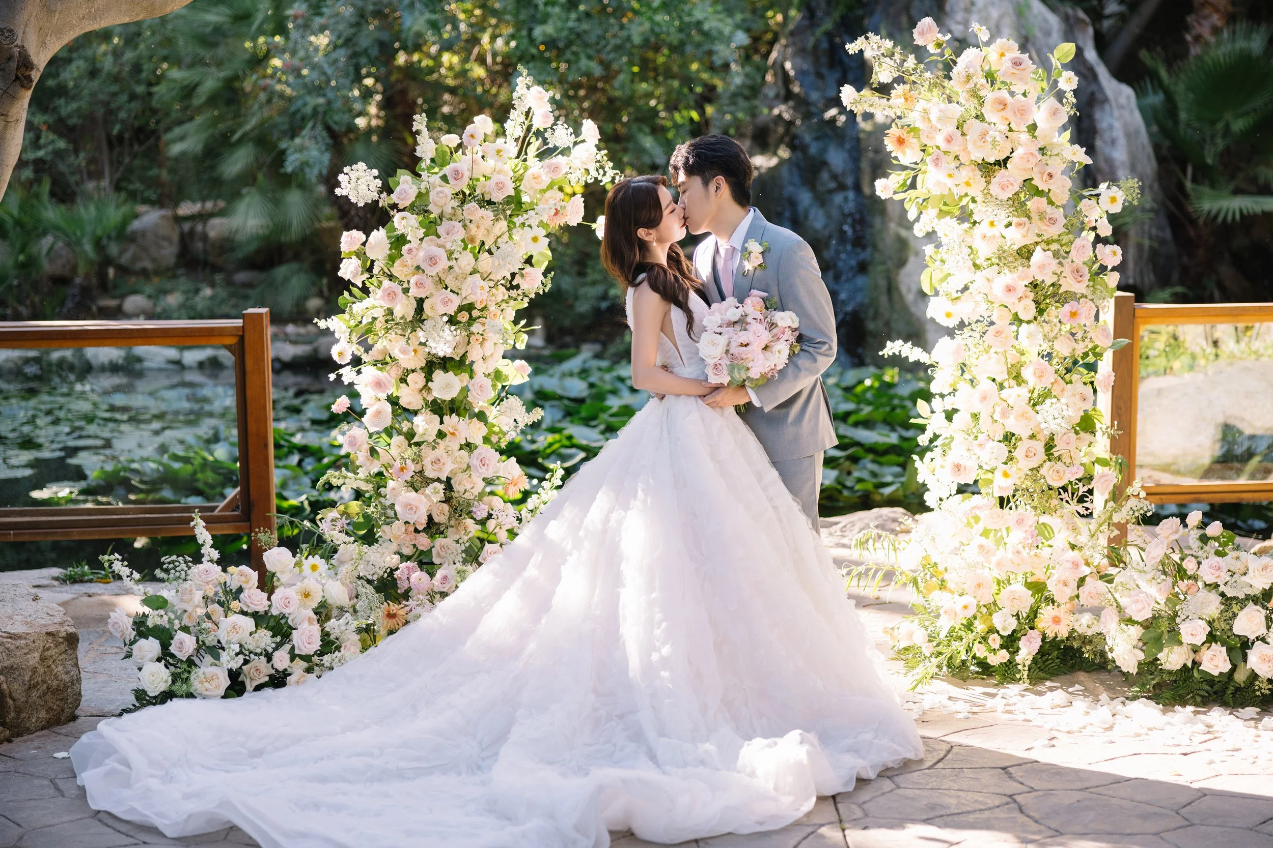 A bride and groom kissing at their outdoor wedding ceremony, surrounded by pink and white floral arrangements and greenery.