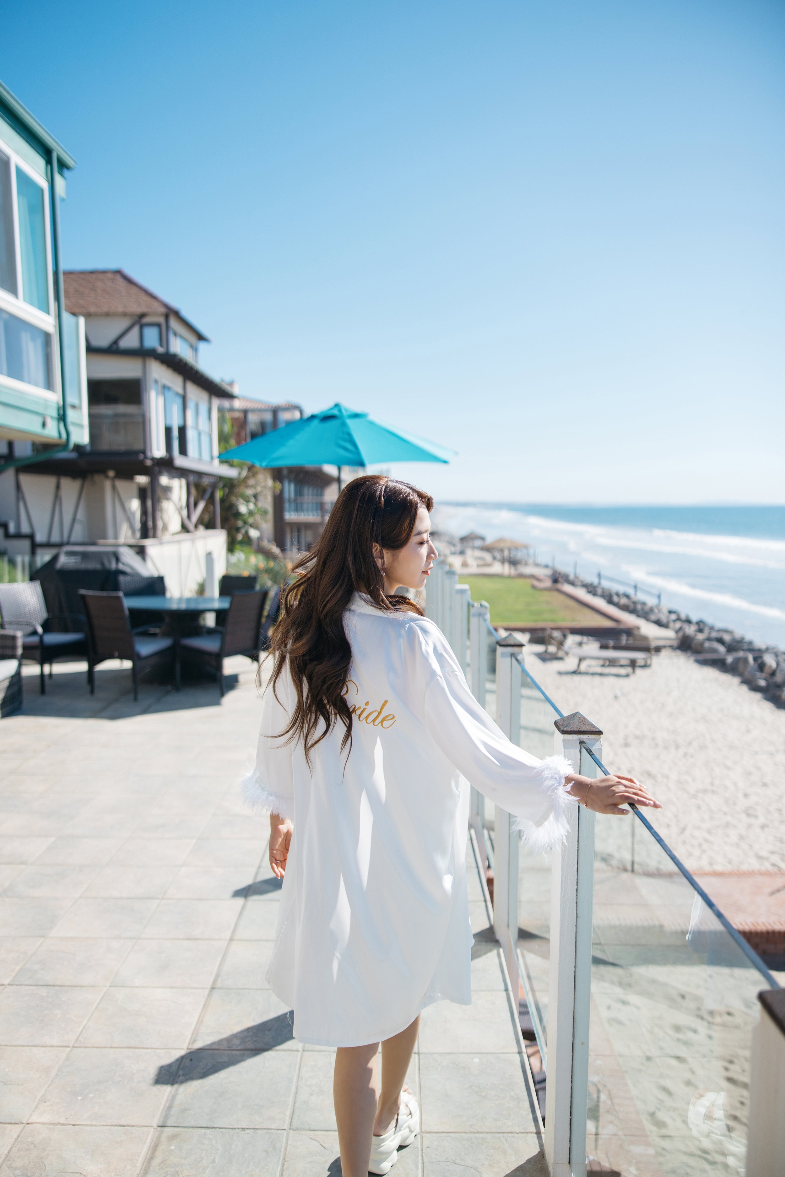 A woman in a white robe with the word 'bride' on the back, standing on a balcony overlooking a beach, with blue sky and ocean in the background.