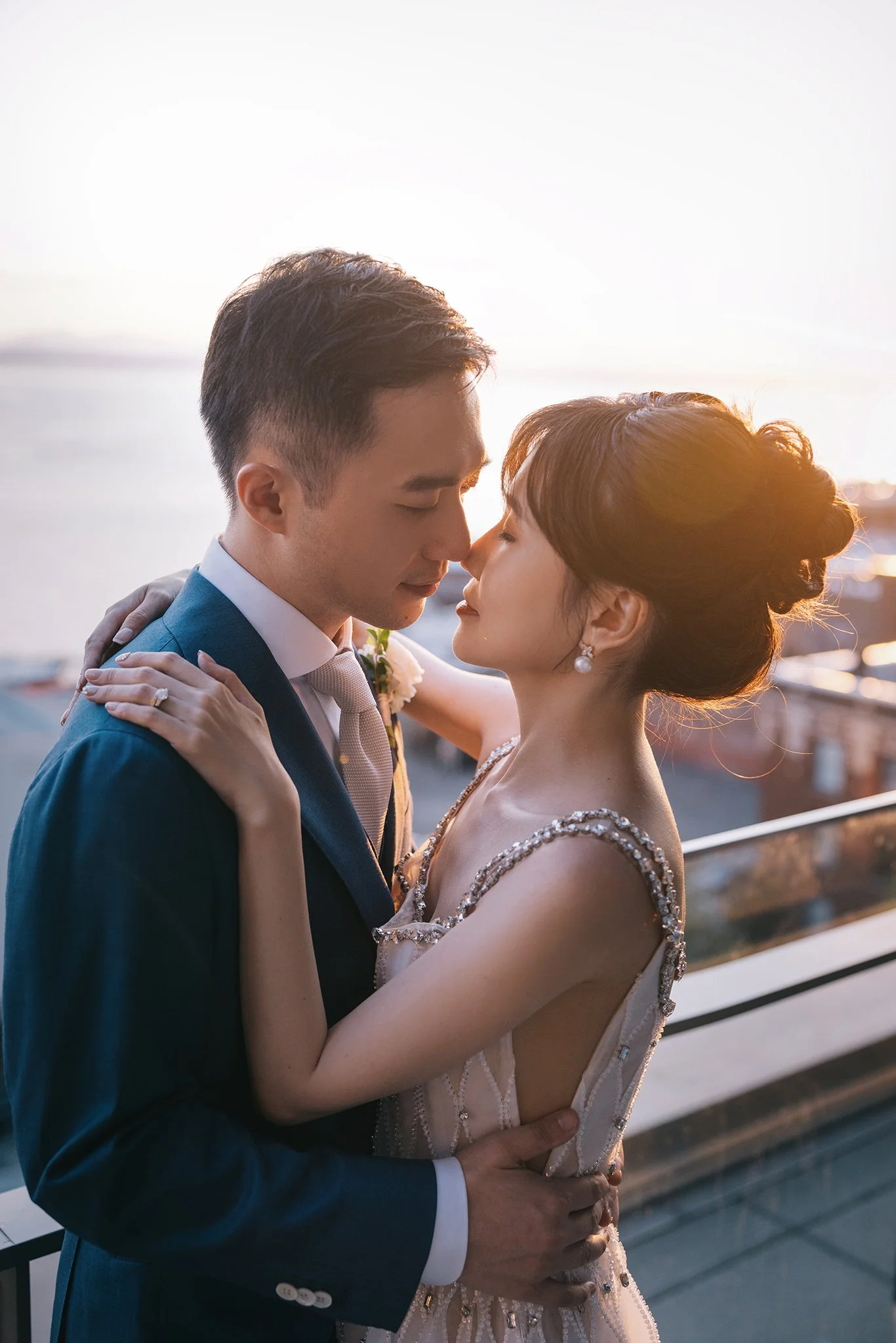 A couple dressed in wedding attire embracing on a balcony at sunset.