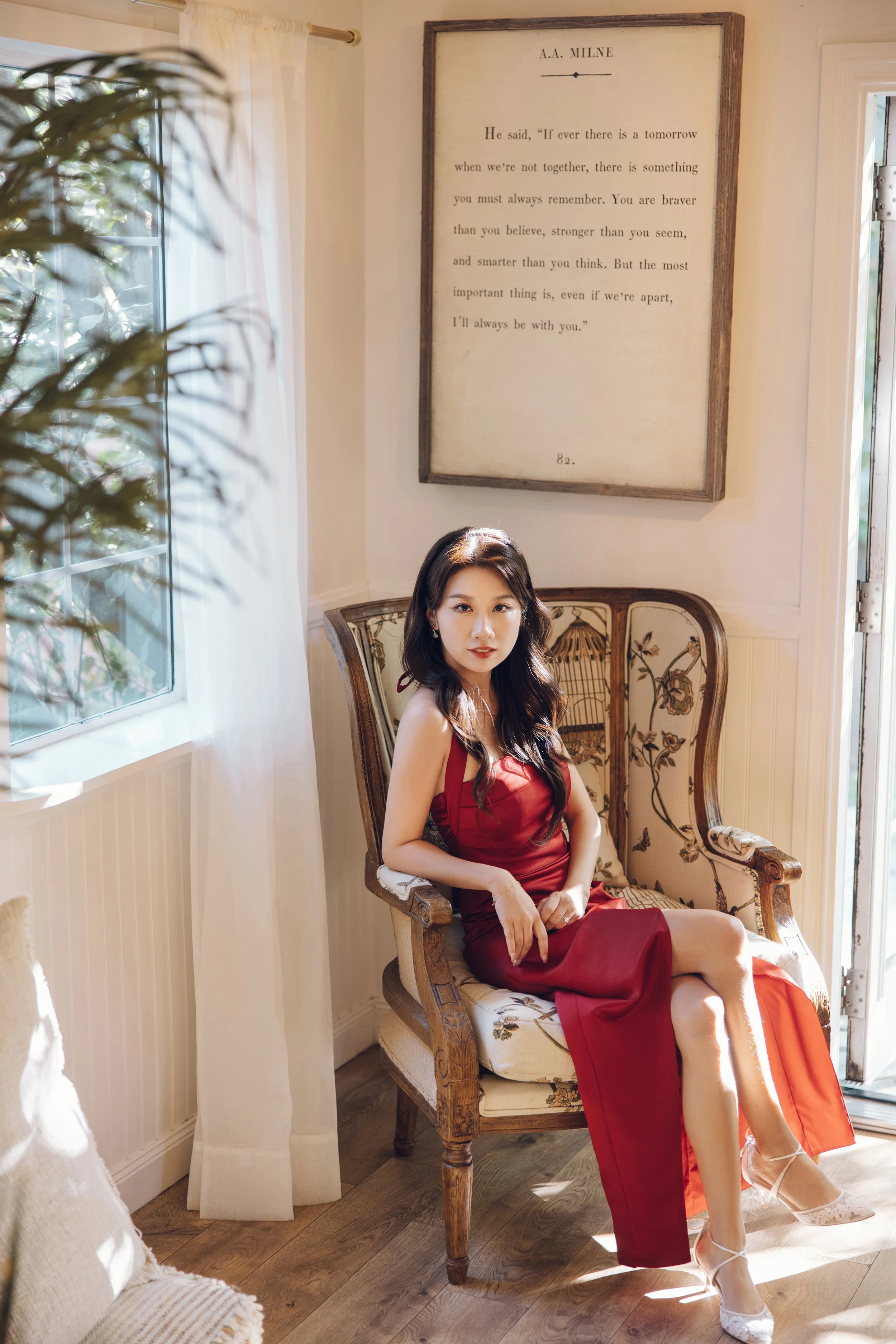 A woman in a red dress sitting on a vintage armchair near a window with sheer curtains, with sunlight illuminating her. There's a framed quote on the wall behind her.