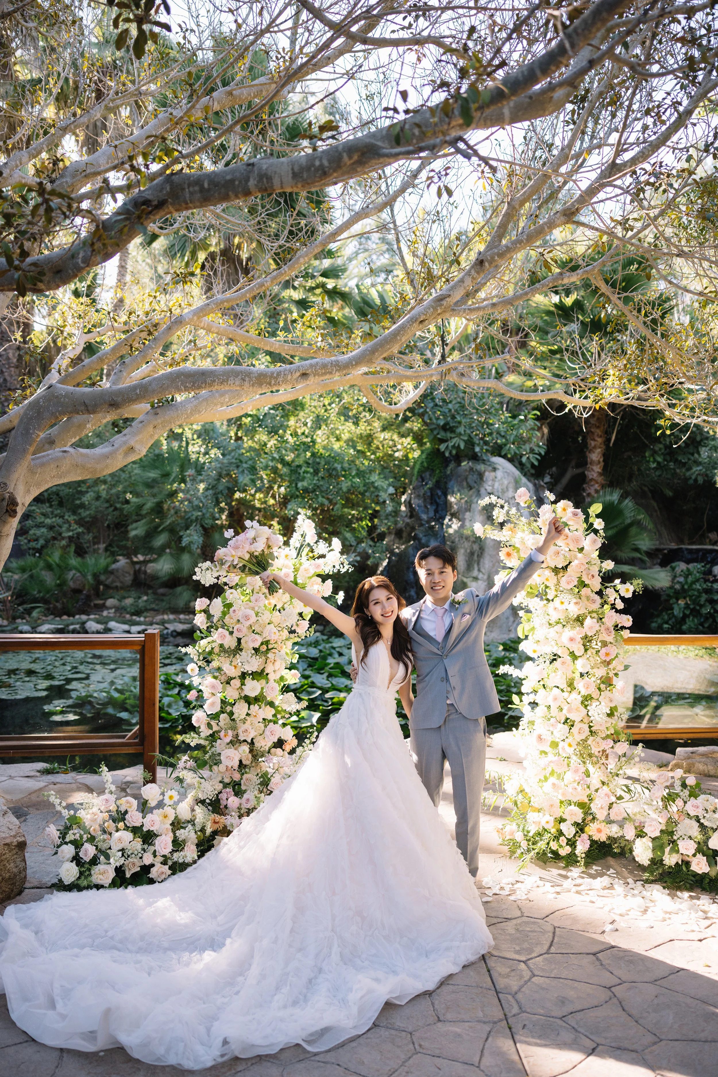 A newlywed couple celebrating outdoors in front of floral arrangements and a pond, with trees and greenery in the background.