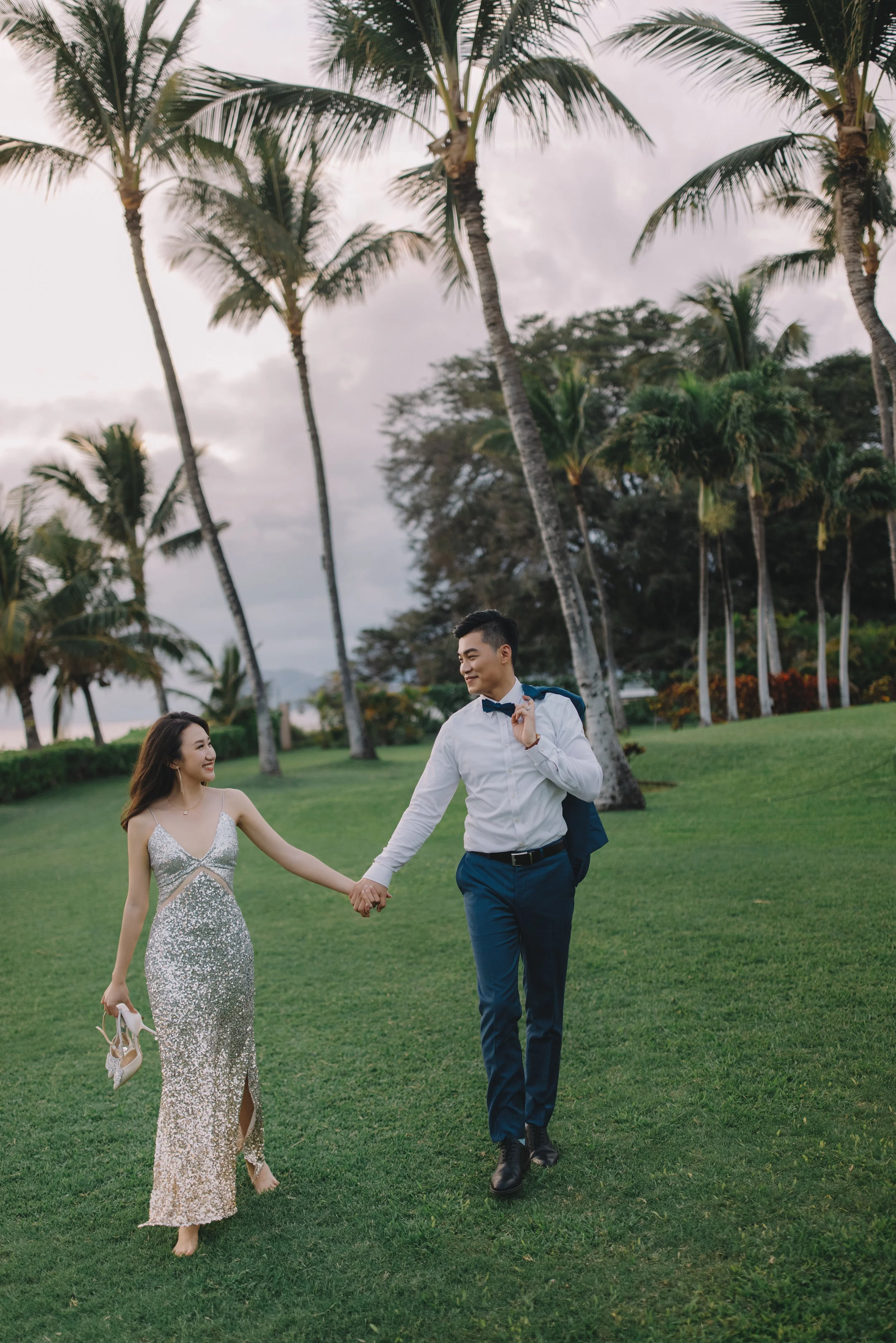 A young couple walking hand-in-hand through a lush green area with palm trees, dressed in formal attire, smiling at each other during an outdoor event or celebration.